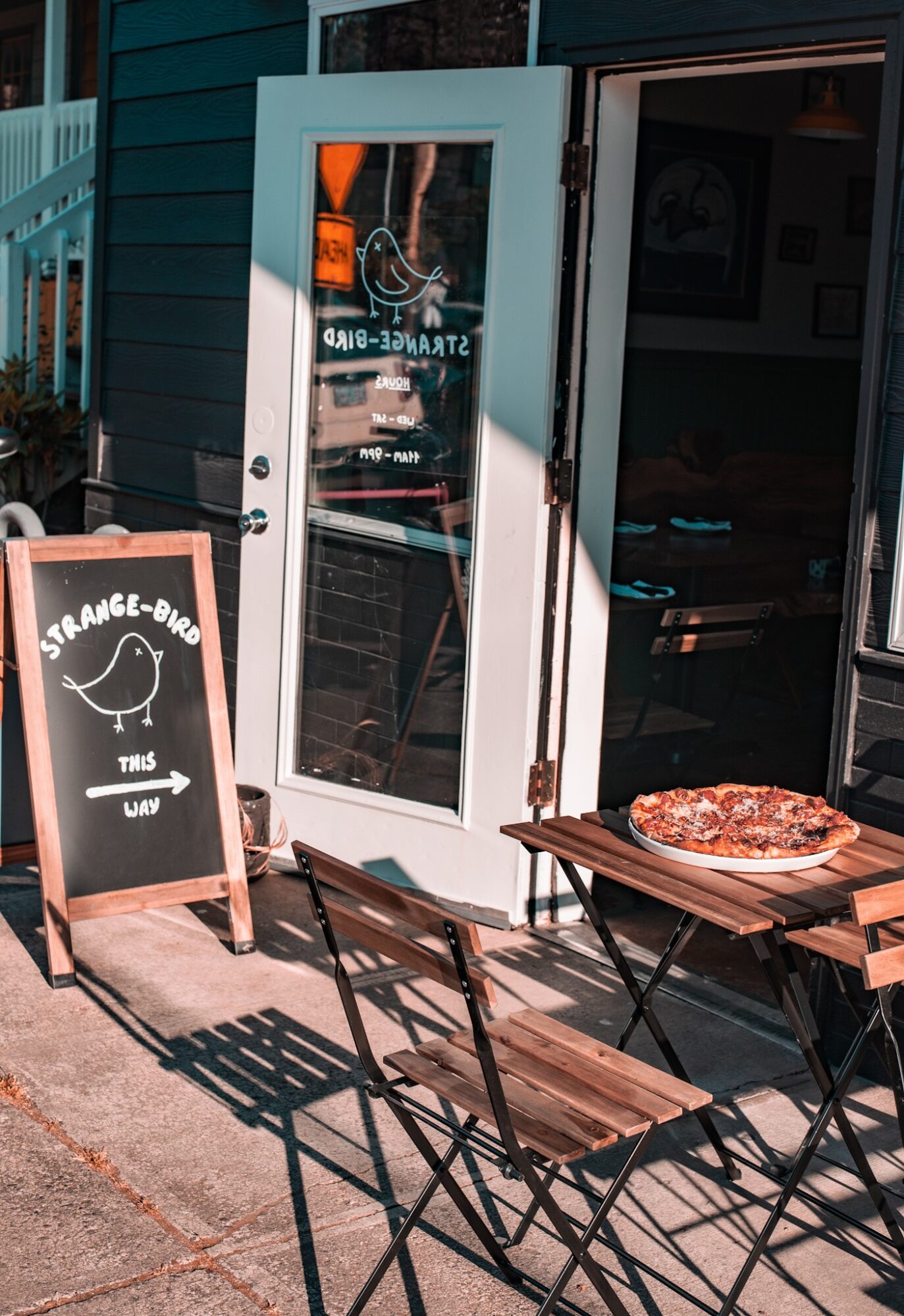 Outdoor seating with a pizza on a table outside a restaurant, with a chalkboard sign and open door, sunlight casting shadows.