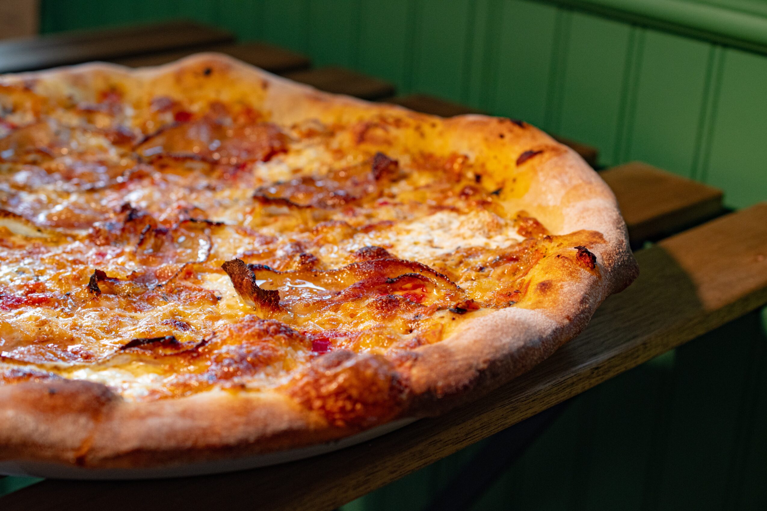 Close-up of a cheese pizza on a wooden serving board, with a green wall in the background.
