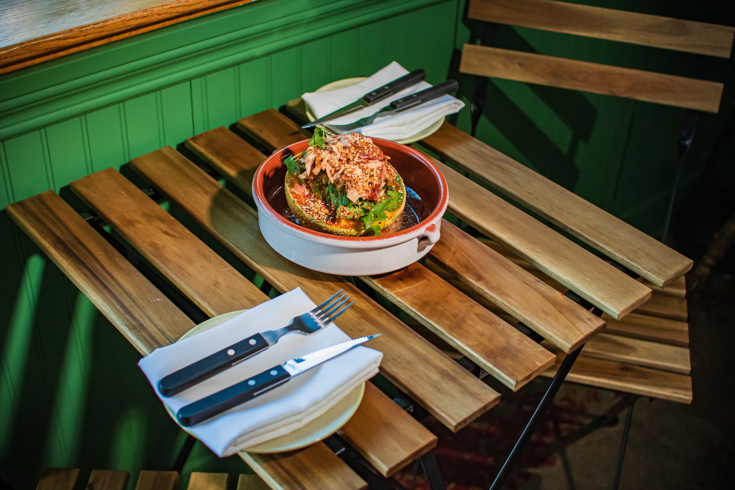 Wooden table with a bowl of food, a plate with utensils, and napkins in a restaurant setting.