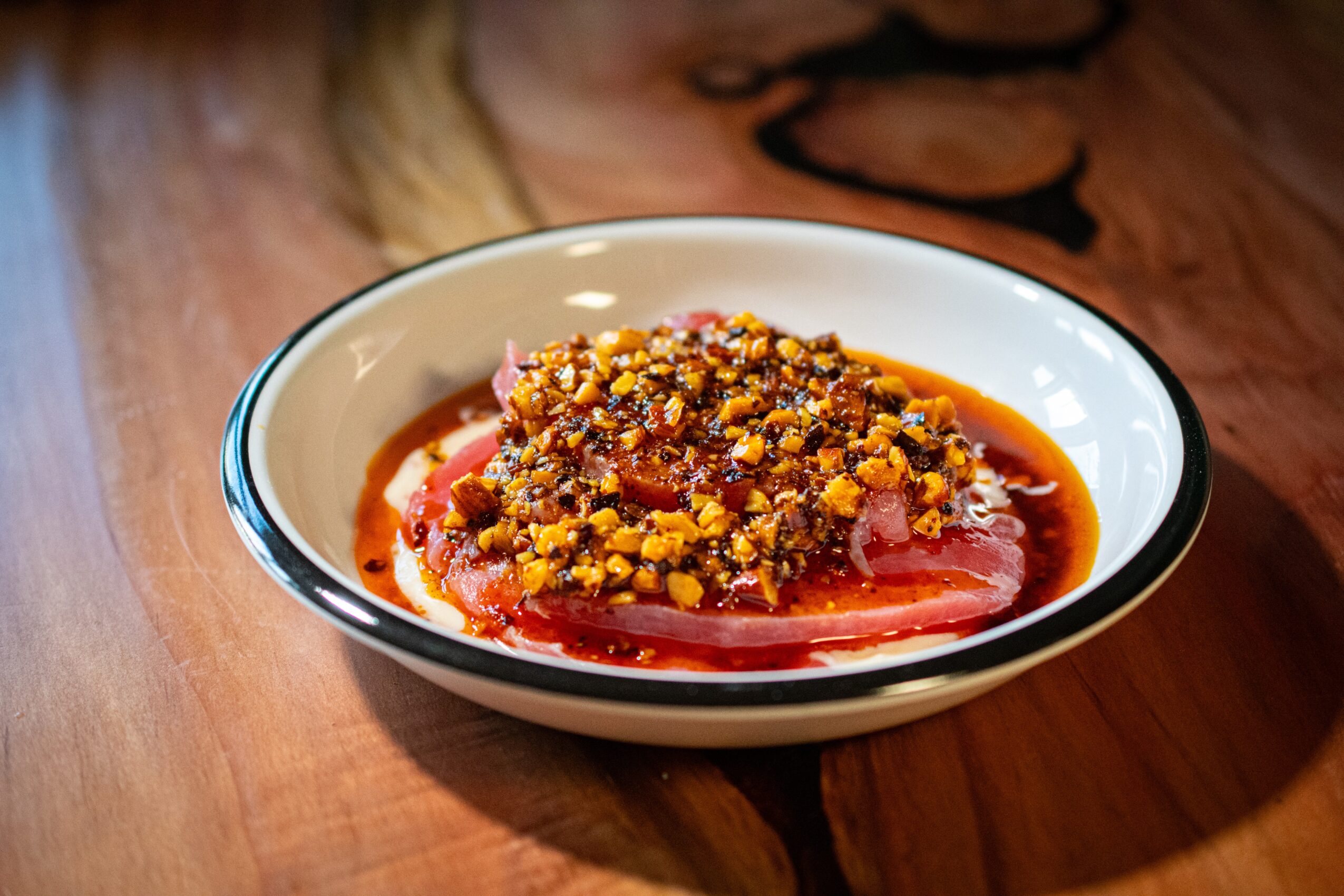 Bowl of food with sauce and toppings on a wooden table, with a blurred background of a person and a plate.