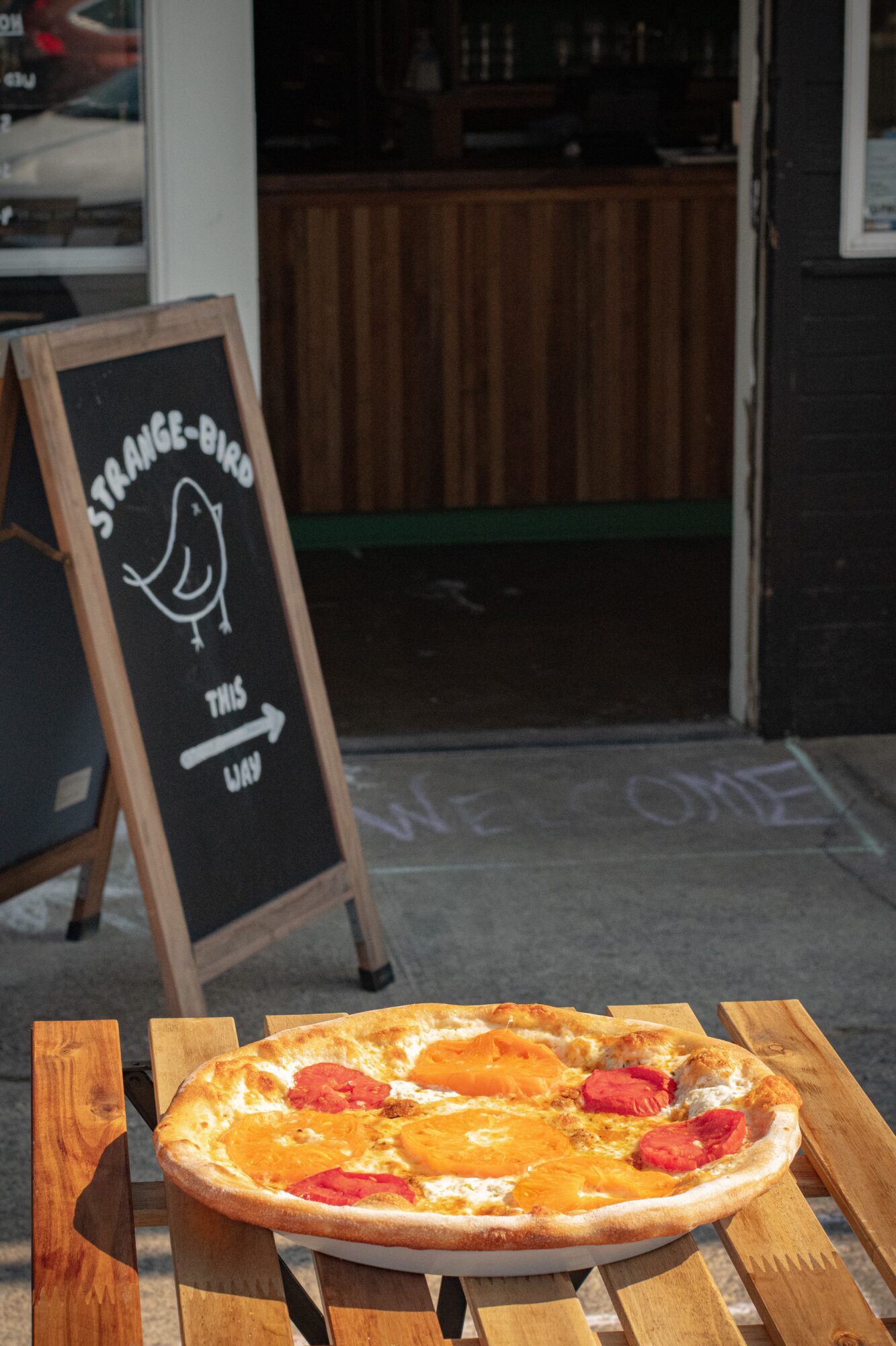Pizza on a wooden table outside a restaurant, with a chalkboard sign nearby. The restaurant interior is visible through the door.