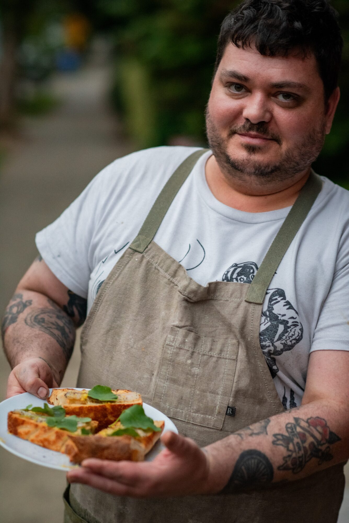 Man with dark hair and beard wearing apron holding a slice of pizza outdoors.