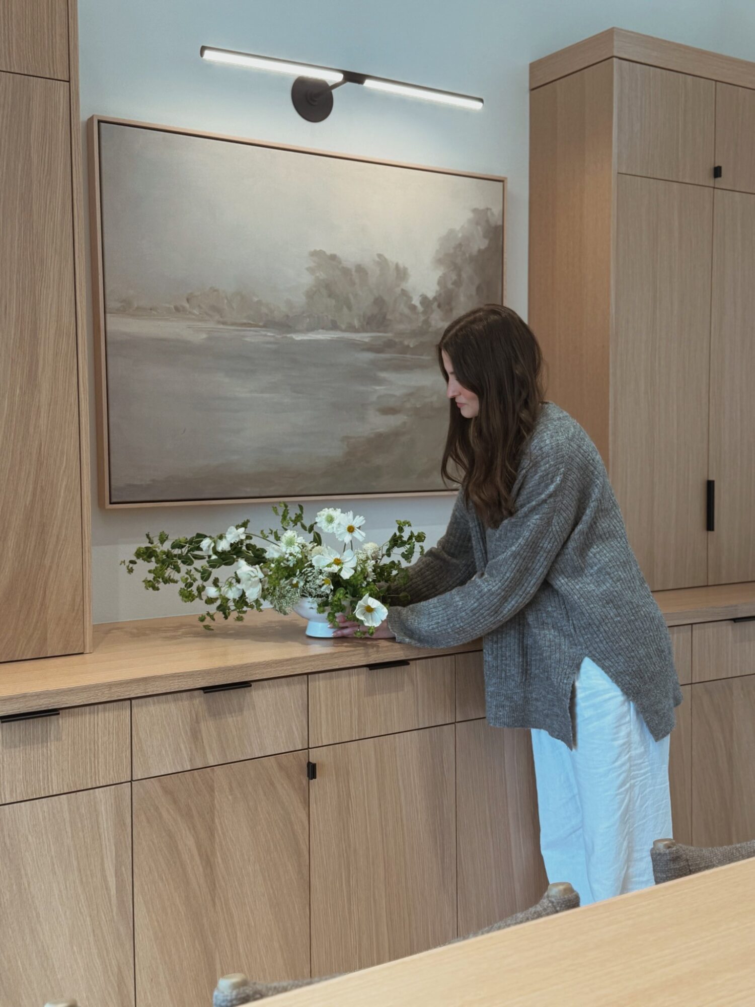 Woman arranging flowers on a wooden cabinet in a room with a large landscape painting and wooden furniture.