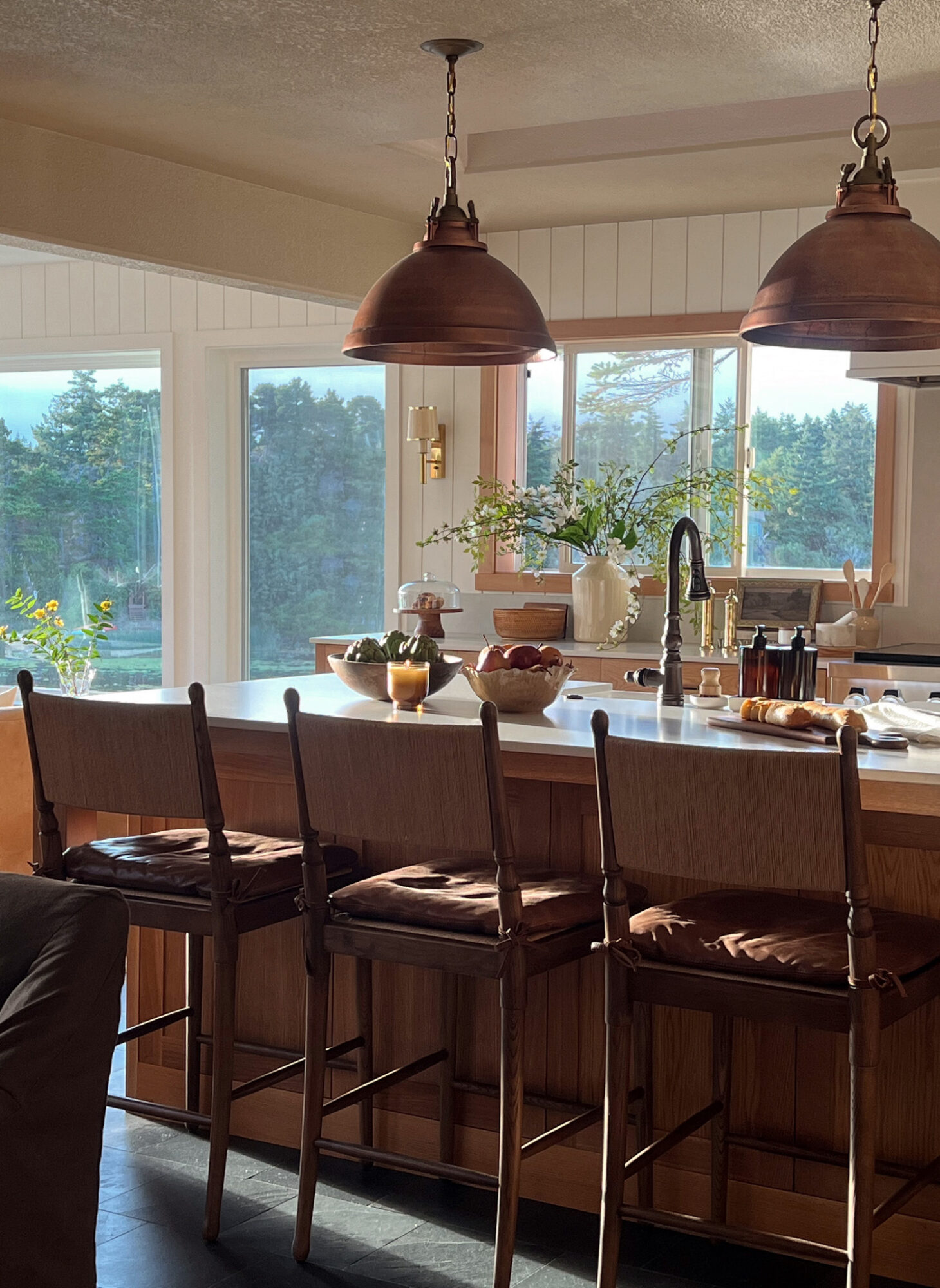 Kitchen island with four chairs, large windows showing outdoor greenery, pendant lights above, and a sink with a faucet.