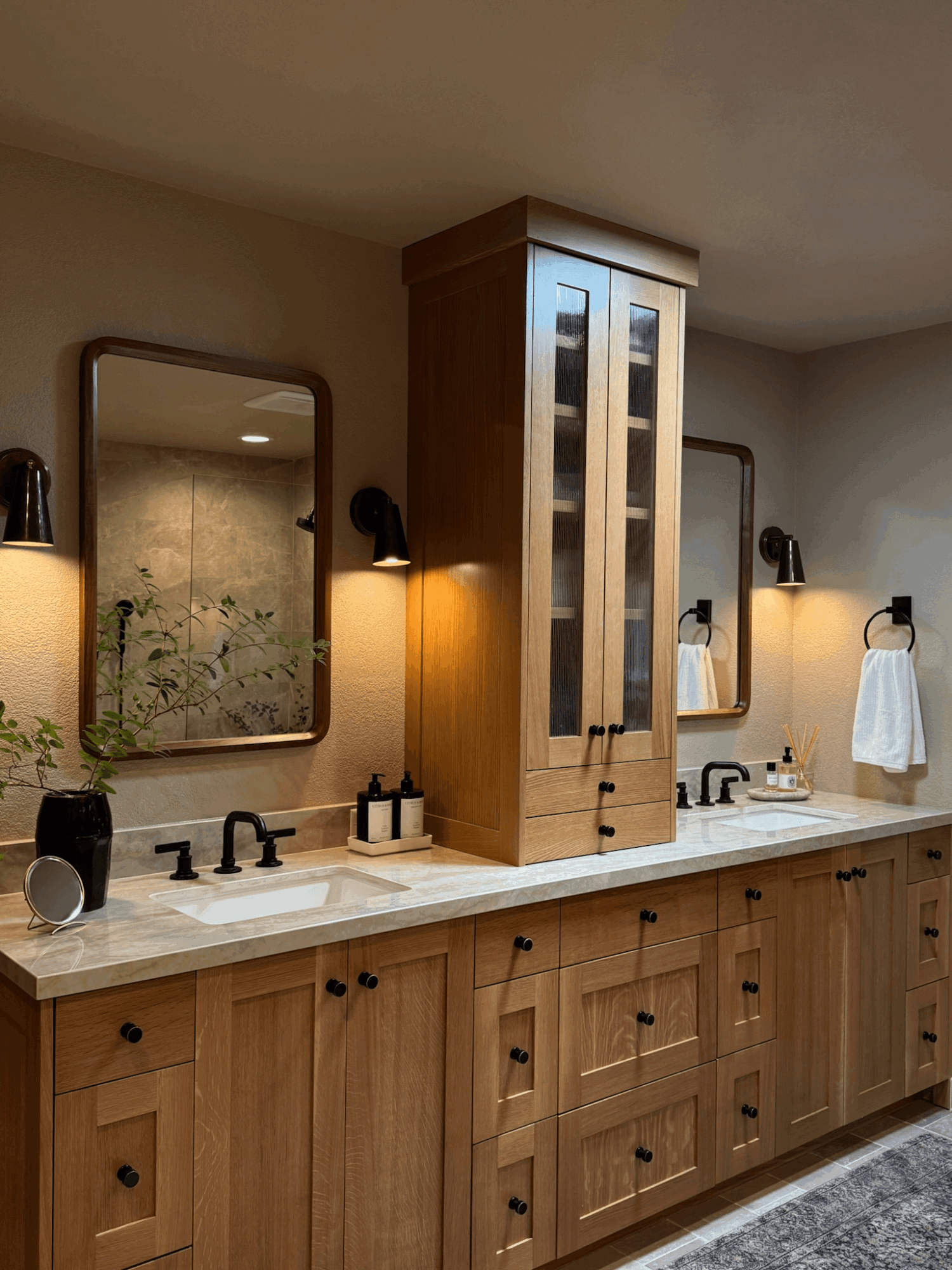 Bathroom vanity with two mirrors, wooden cabinets, and a tall central cabinet, with sinks and black fixtures, illuminated by wall lights.