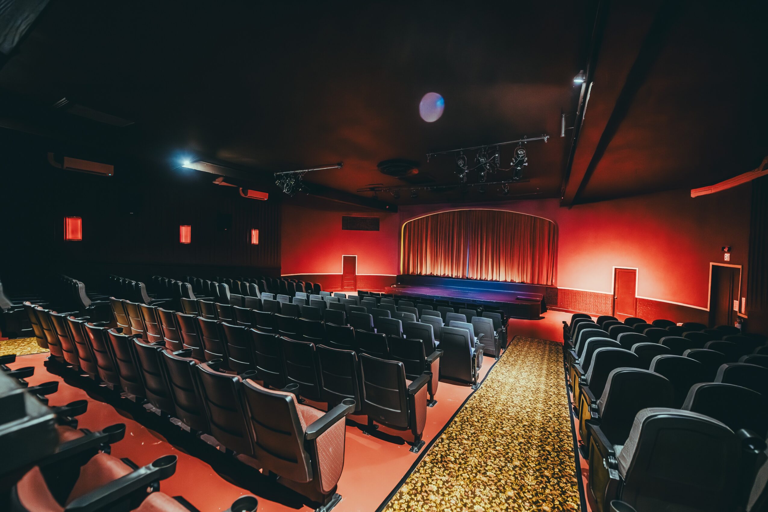Empty theater with rows of seats facing a stage with red curtains, dim lighting, and a patterned carpet aisle.