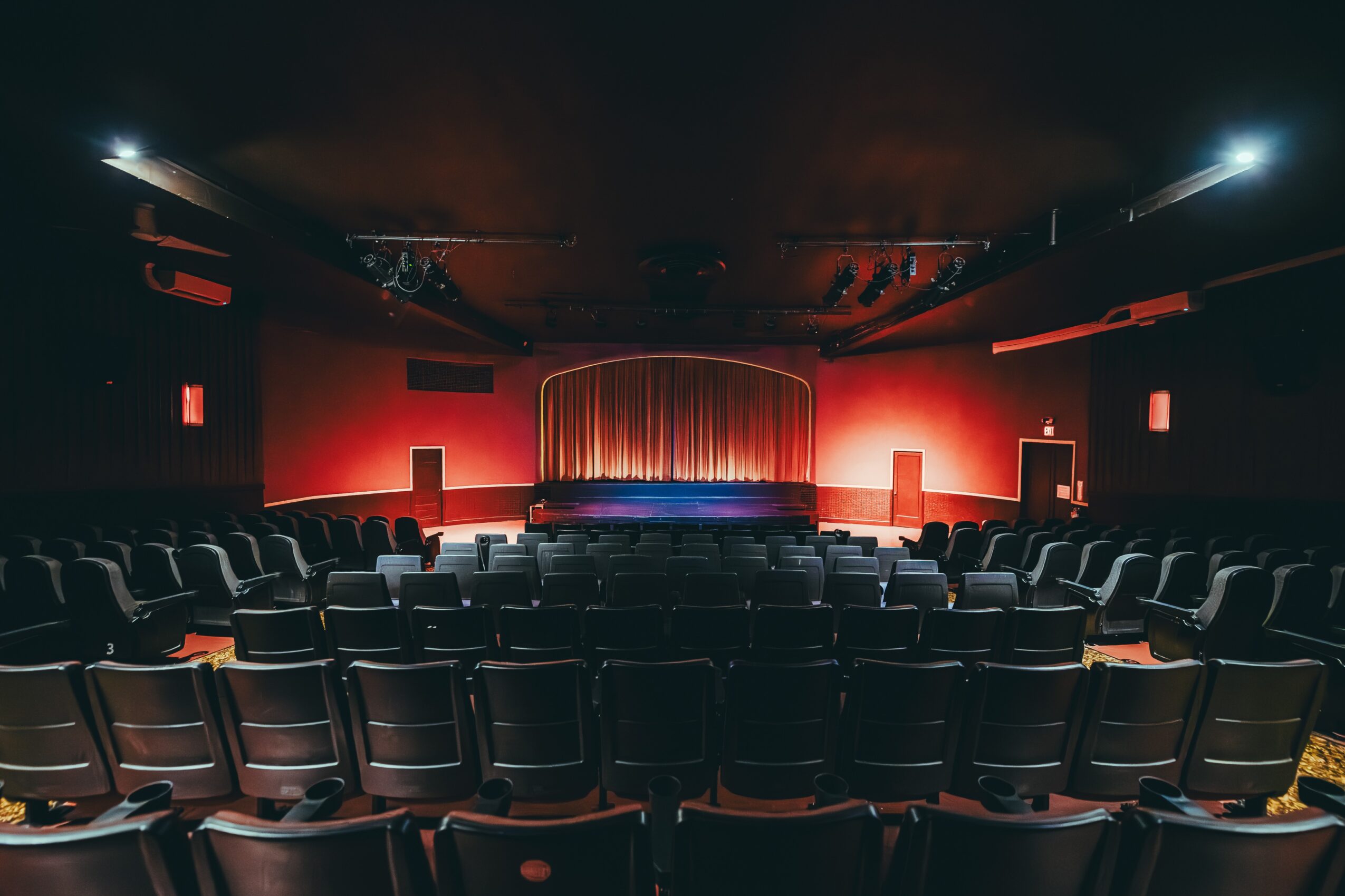 Empty theater with a stage, red walls, and rows of seats facing the stage, dim lighting, and a curtain on stage.