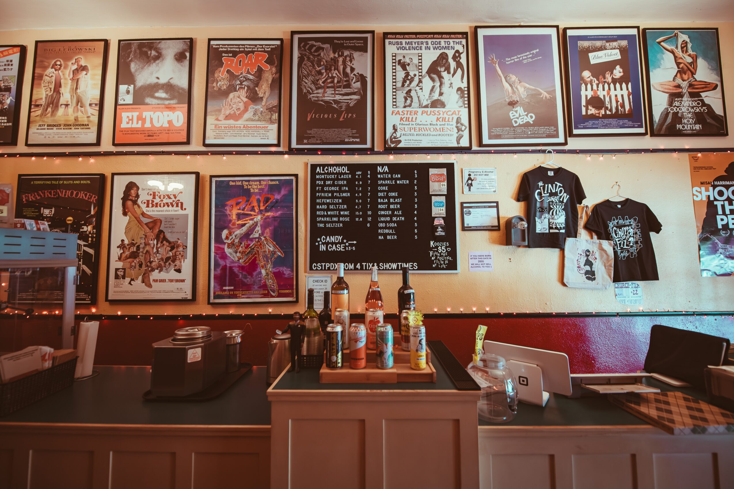 Bar counter with bottles, posters on the wall, and t-shirts displayed, in a dimly lit setting.