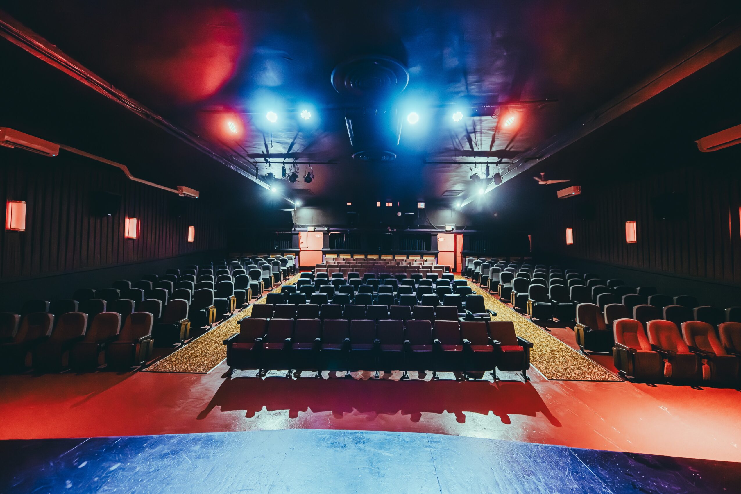 Empty auditorium with rows of chairs facing a stage, illuminated by blue and red lights, with a dark ceiling and walls.
