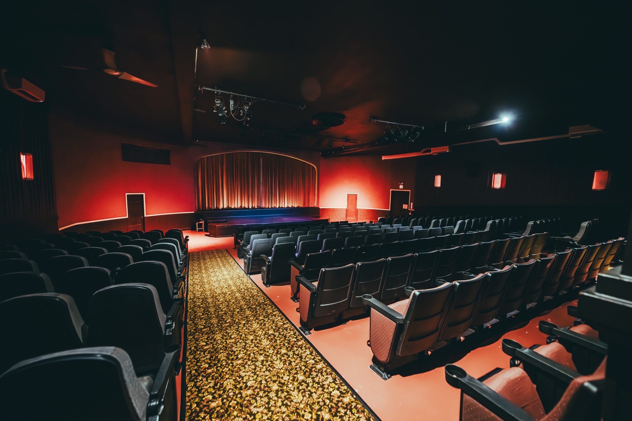 Empty theater with rows of seats facing a stage with red curtains, dim lighting, and a patterned carpet aisle.