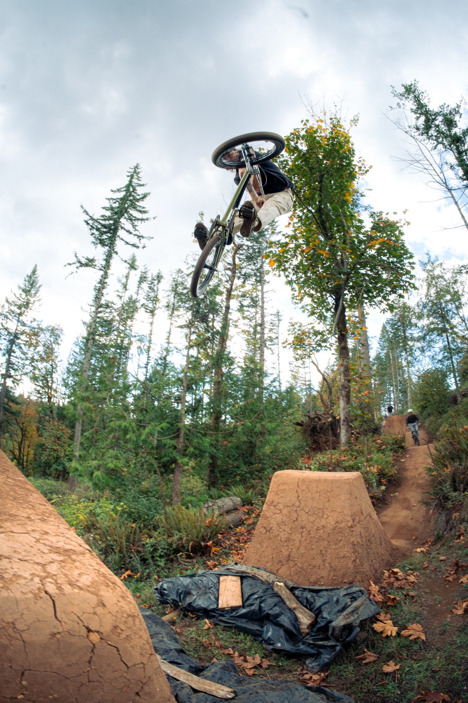 Bicycle airborne above dirt ramps in a forested area with trees and cloudy sky.
