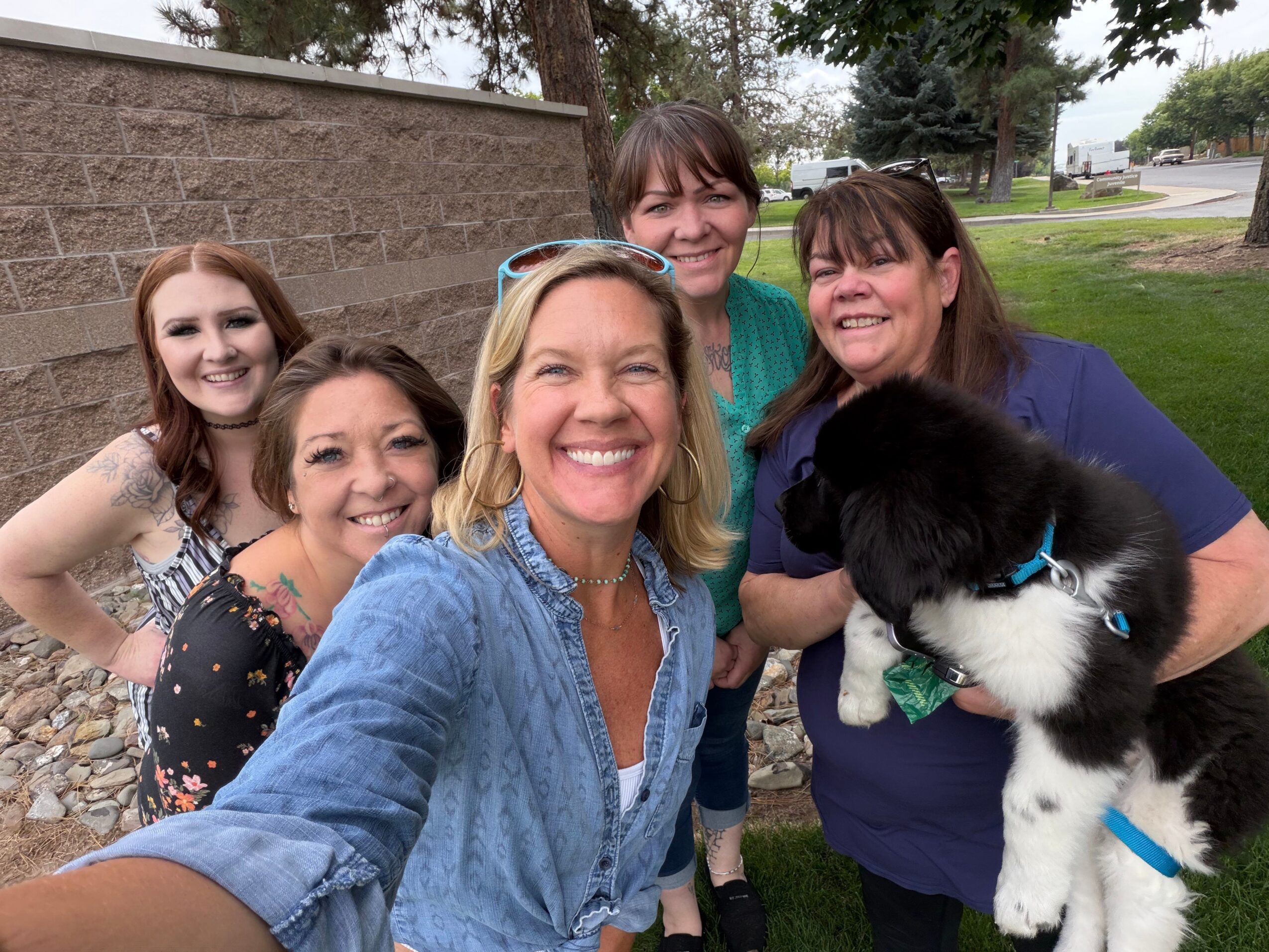Four women and a puppy outdoors, smiling and posing for a photo, with trees and a stone wall in the background.