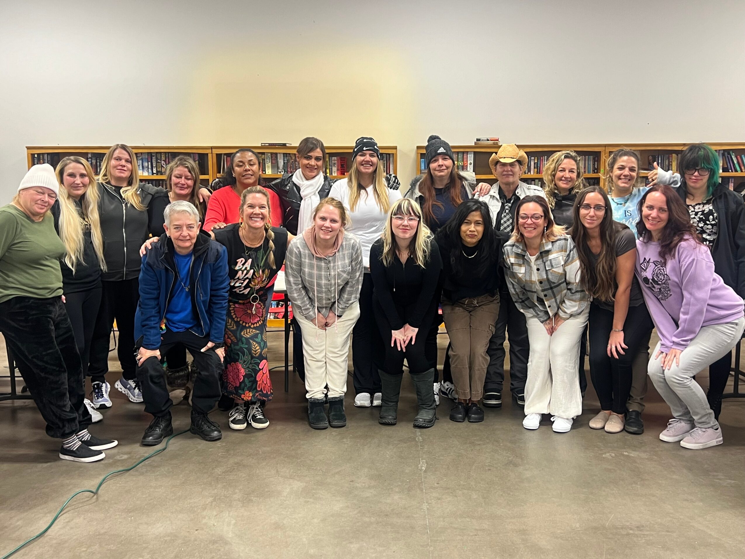 Group of 20 people standing indoors, smiling, some wearing casual clothing and hats, in a room with bookshelves.