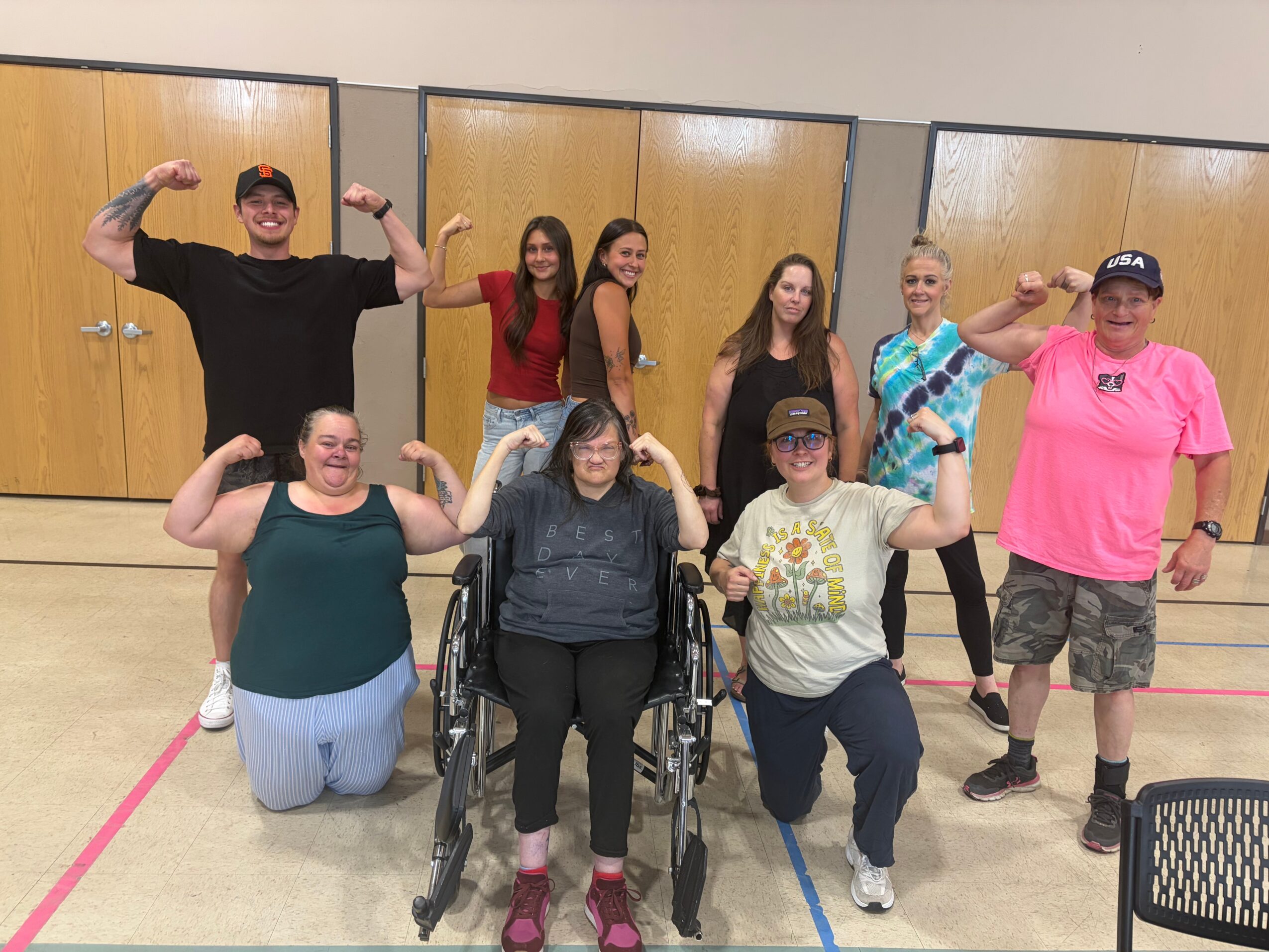 Group of nine people, some in wheelchairs, posing indoors with arms flexed, smiling, and making fists, in a gym or activity room.