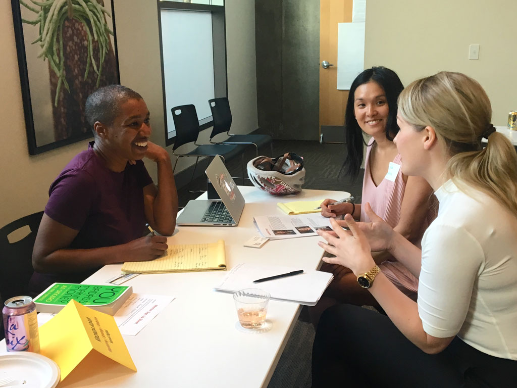 Three women sit at a table in a meeting room, smiling and engaging in conversation with laptops and papers.