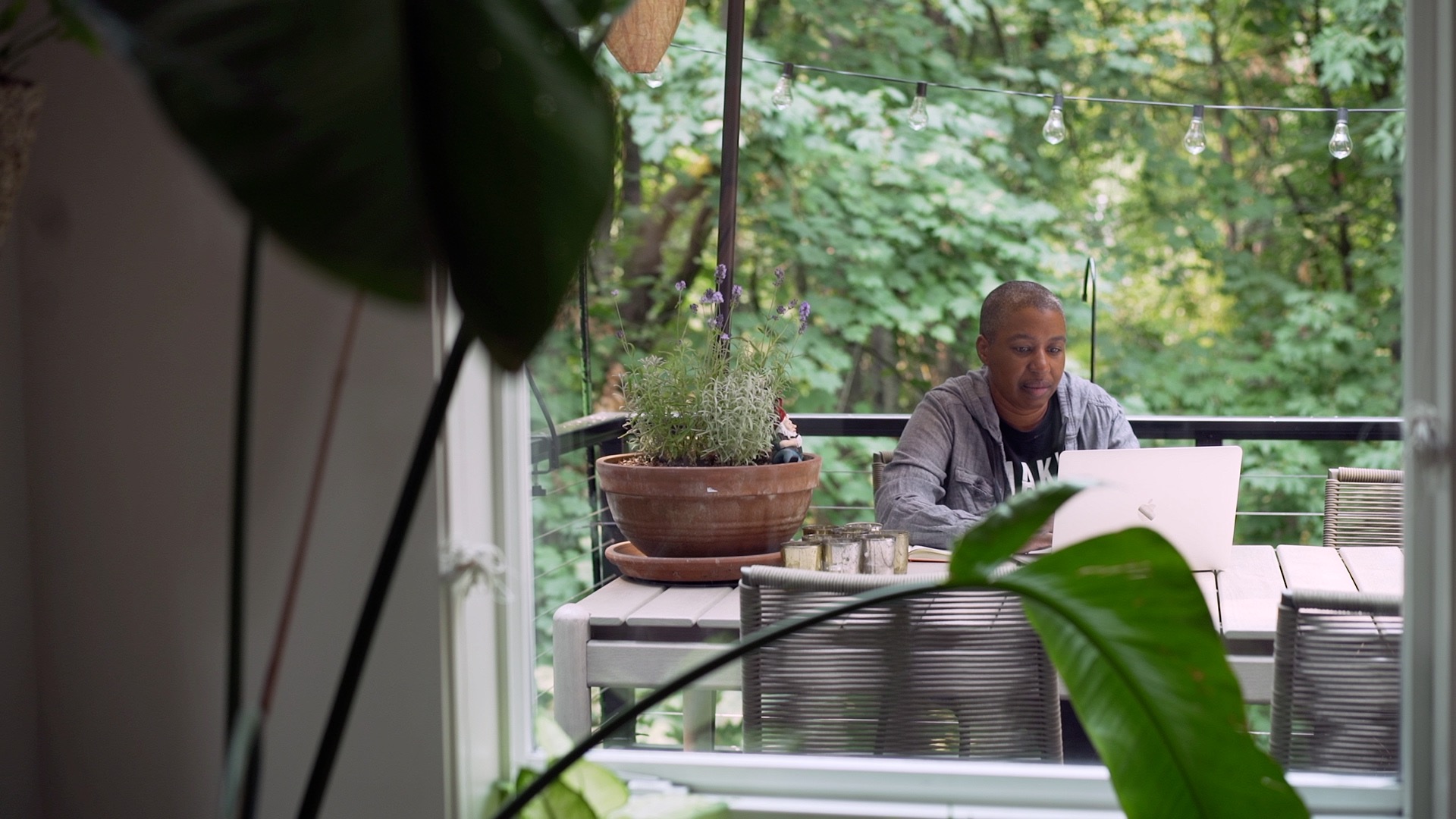Person working on a laptop at a table in a bright room with large windows and greenery outside.