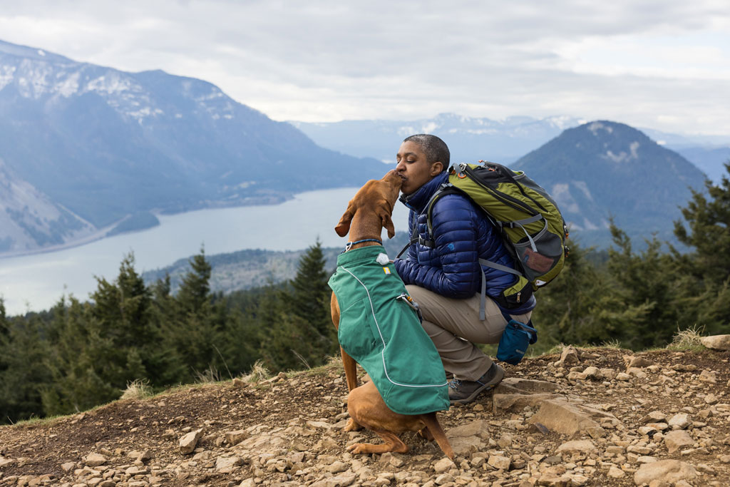 Person hugging a dog outdoors with mountains and a lake in the background.