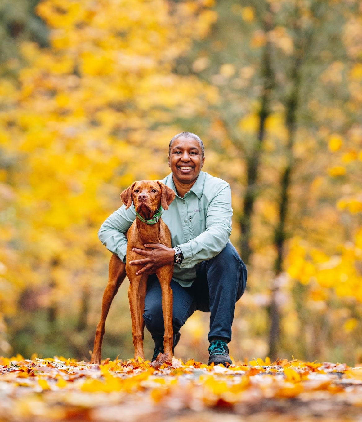 Person crouching outdoors with a brown dog in a park with autumn leaves, smiling happily, surrounded by yellow and orange trees.