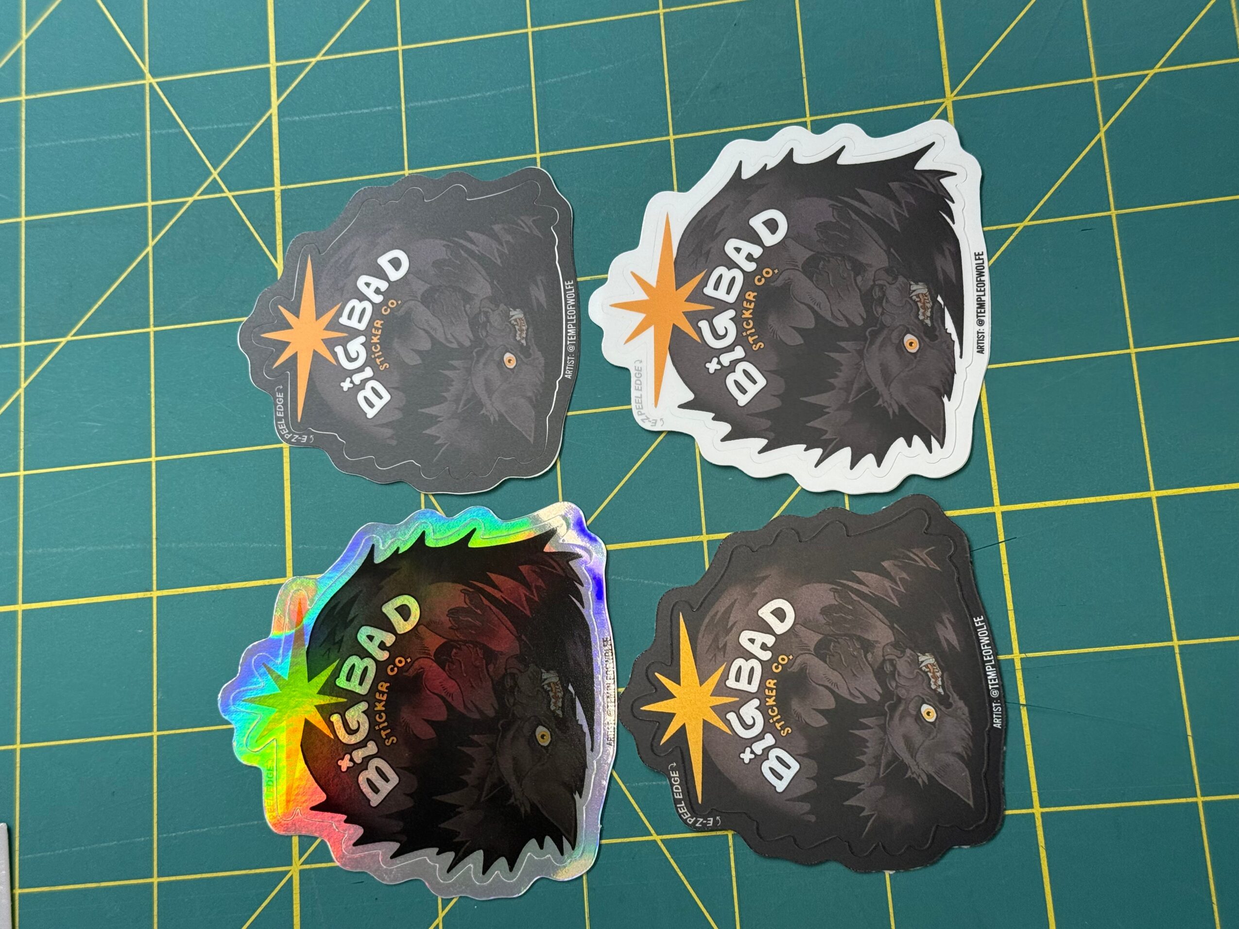 Four stickers of a black dog with a star and text, arranged on a green cutting mat, one sticker has rainbow colors.