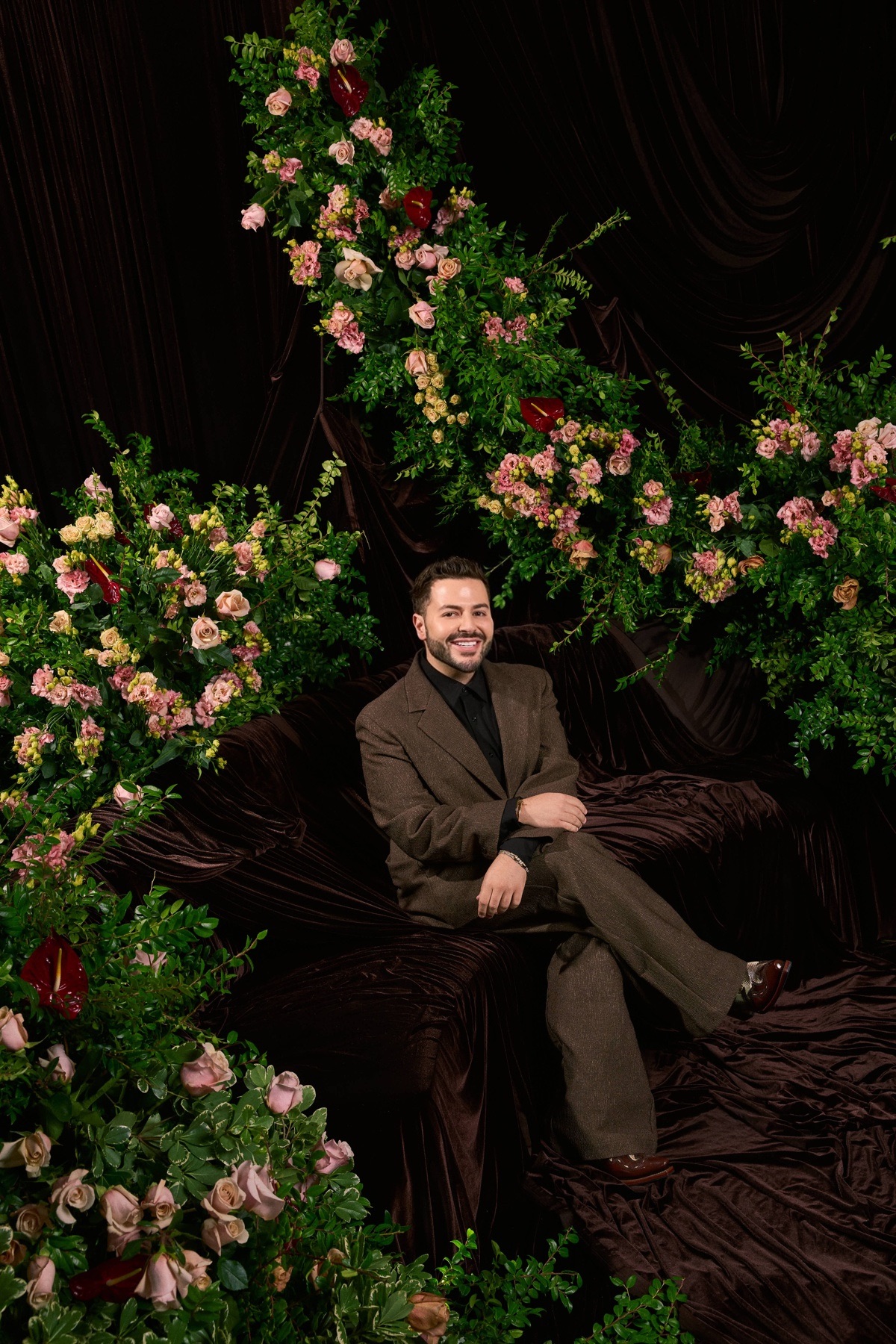 Man sitting on a dark velvet sofa surrounded by pink and green floral arrangements, smiling, with dark drapery background.