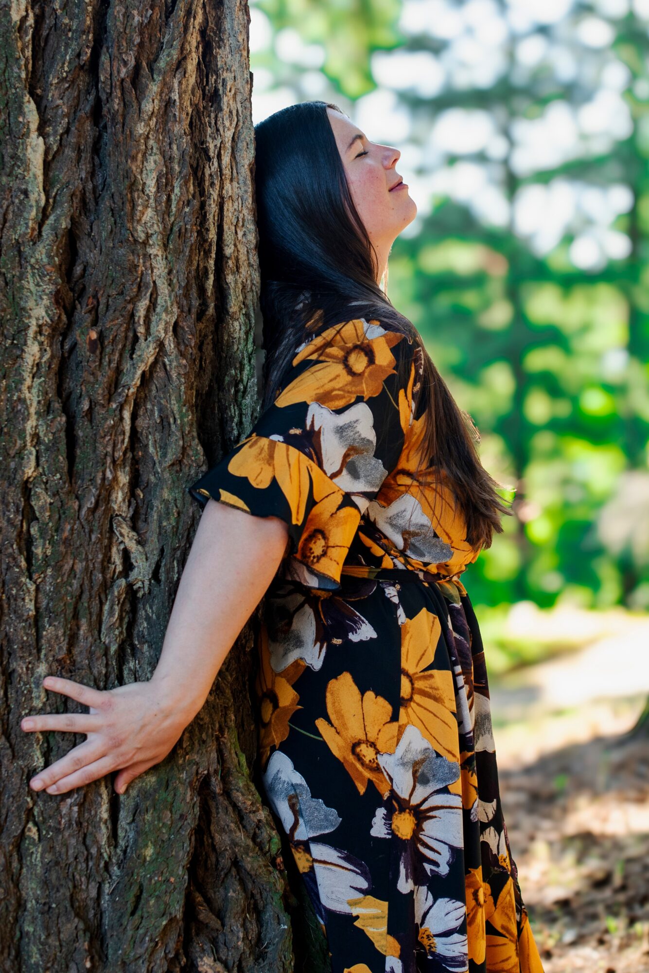 Woman leaning against a tree outdoors with eyes closed, wearing a floral dress, surrounded by green trees.