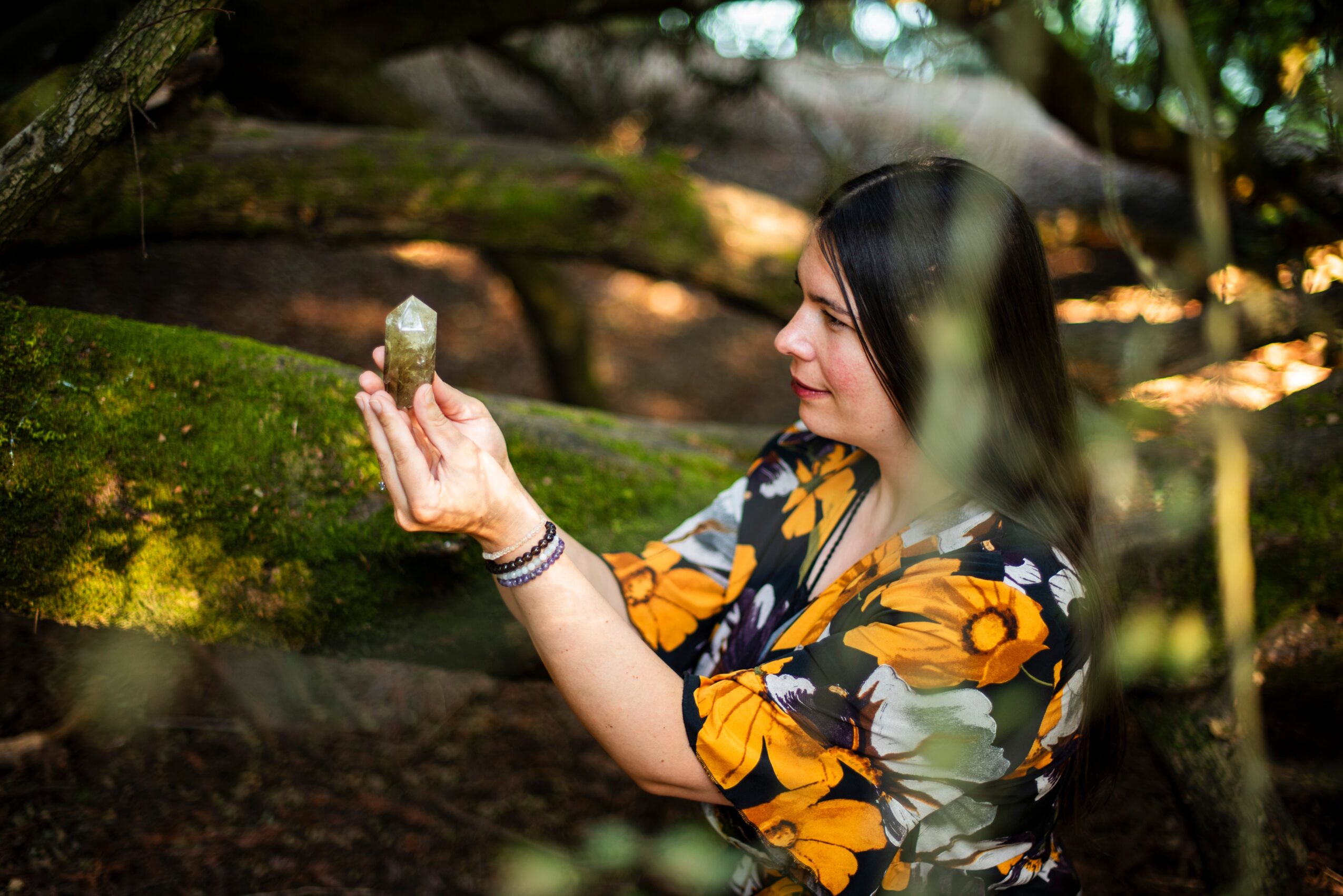 Woman with long dark hair wearing a floral dress, holding a small object in a forest setting.