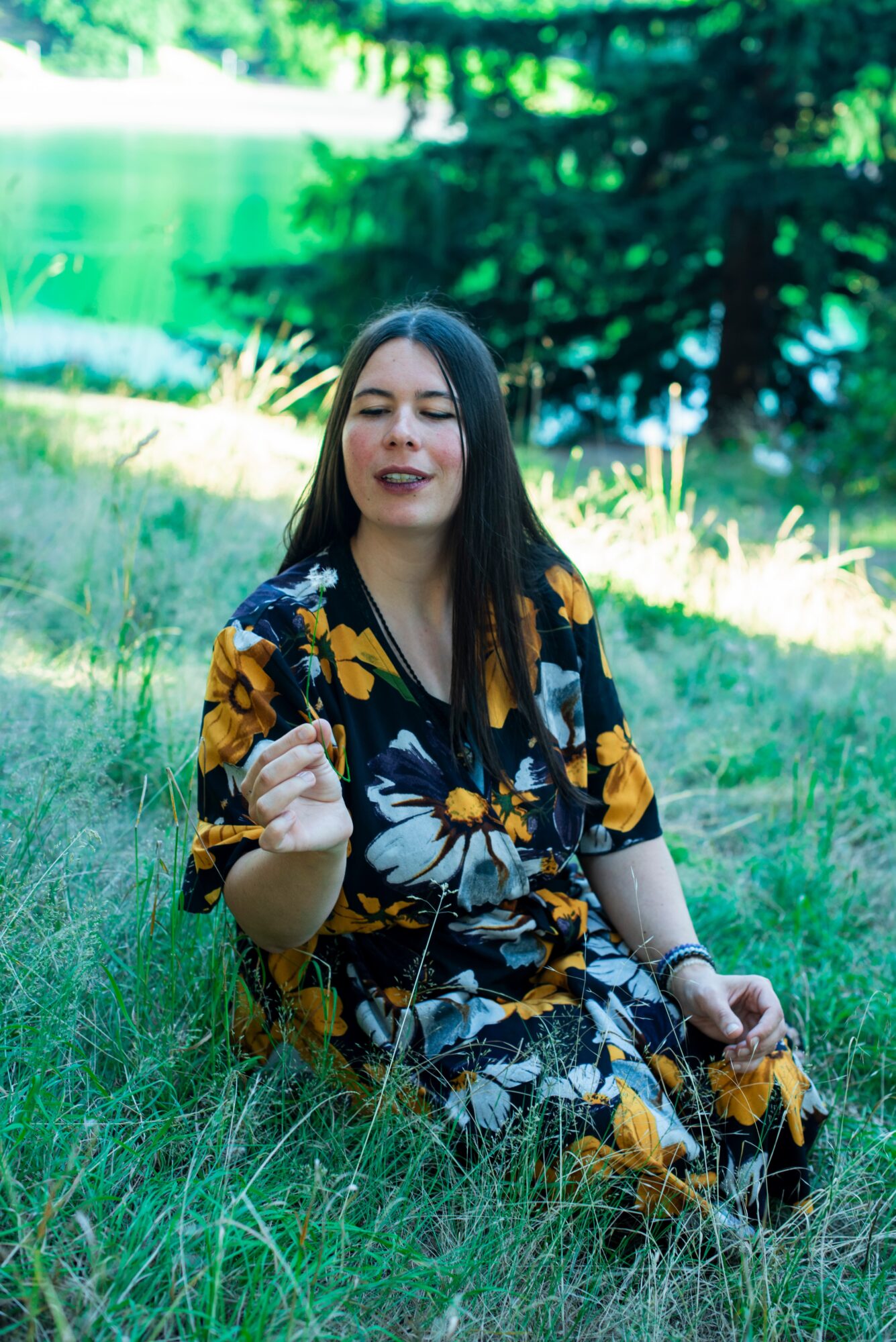Young woman with long dark hair sitting on grass near a lake, wearing a floral dress, smiling with eyes closed.