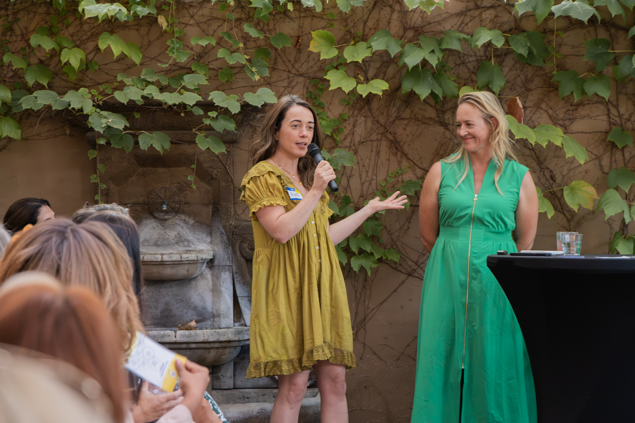 Two women speaking to an audience outdoors, one holding a microphone, surrounded by people, with a vine-covered wall behind.