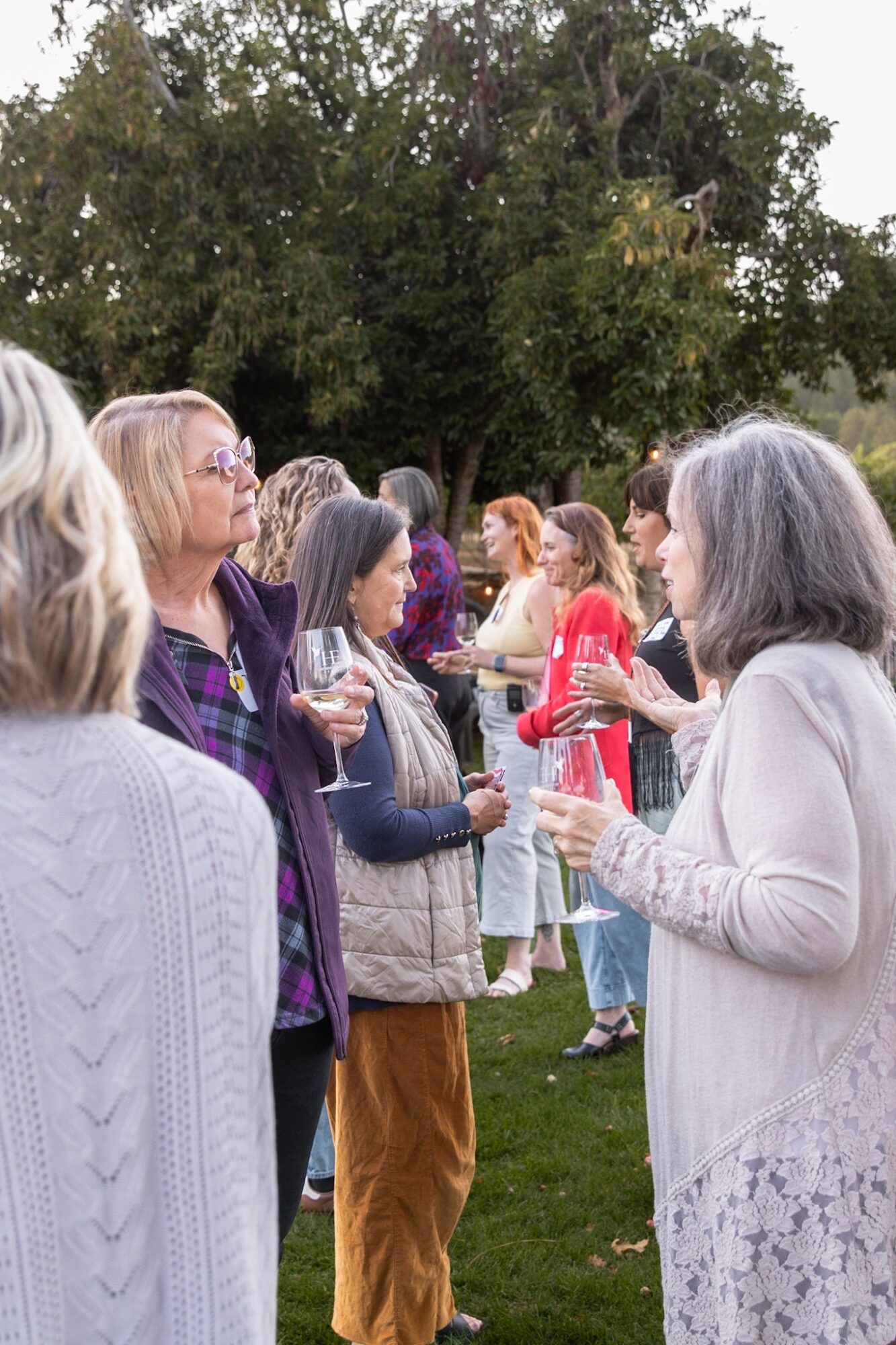 Group of women standing outdoors, talking and holding drinks, with trees in the background.