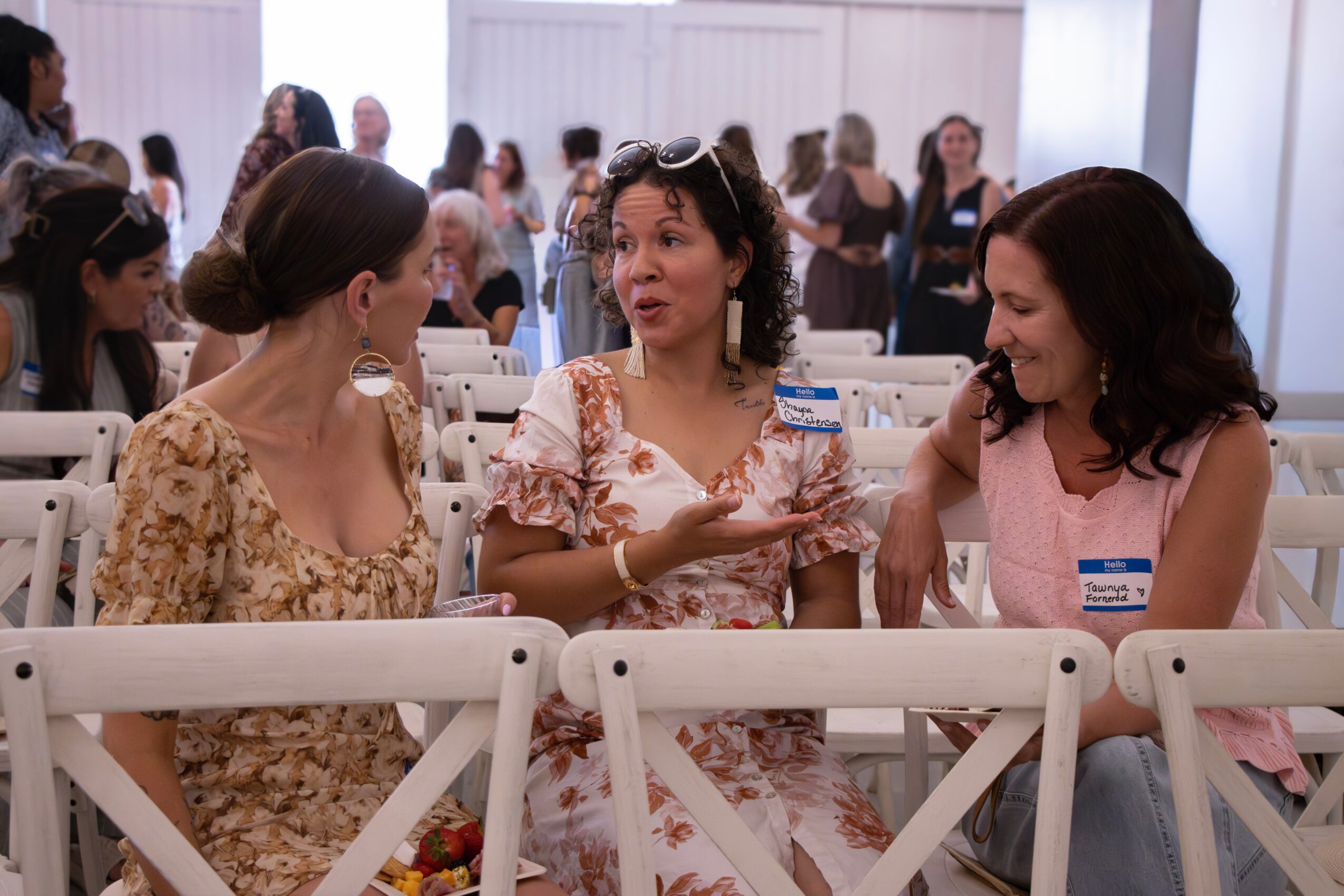 Three women sitting and talking in a bright room with many people in the background.