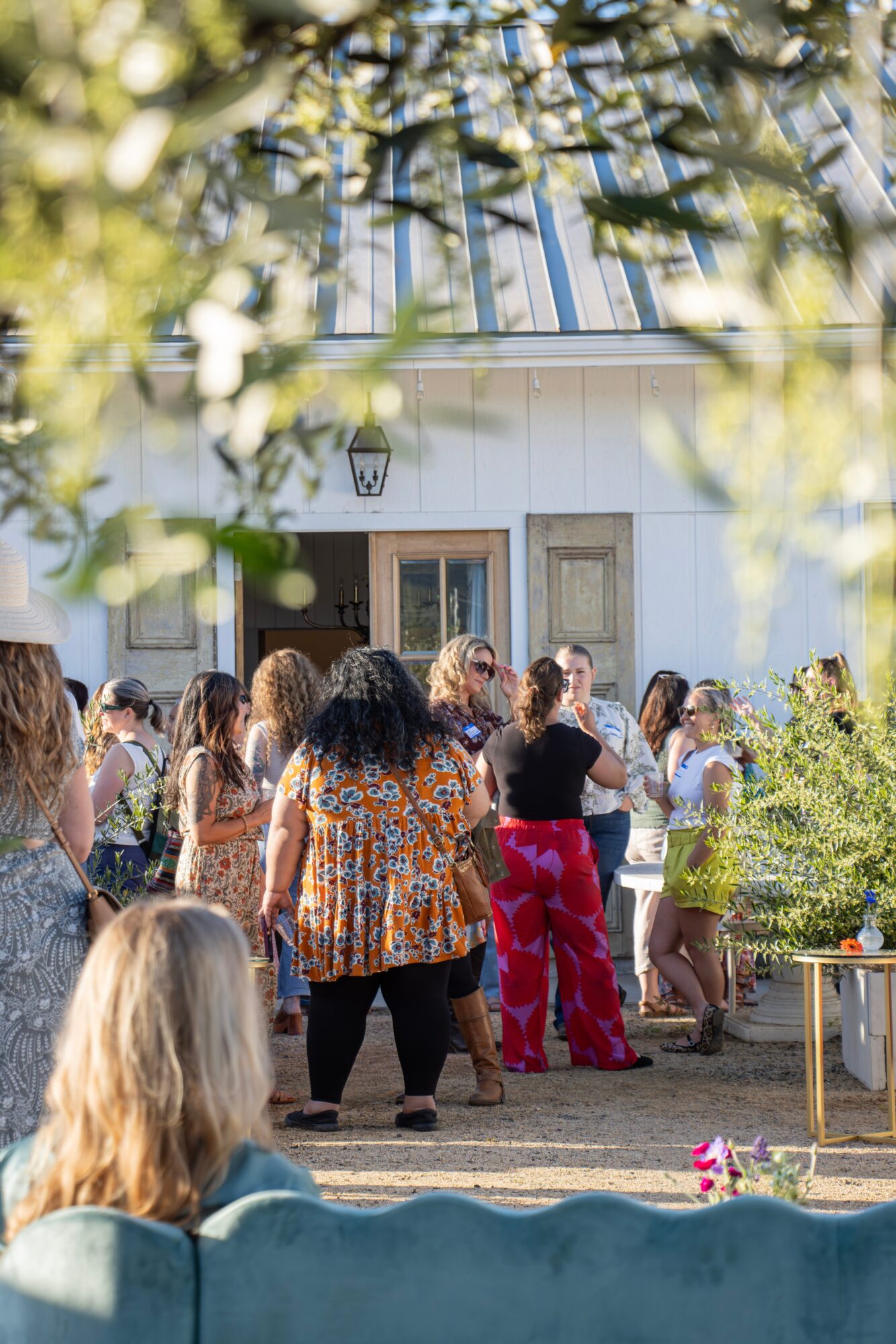 Group of people gathered outside a white building with a metal roof, some dancing and socializing, with trees and sunlight.