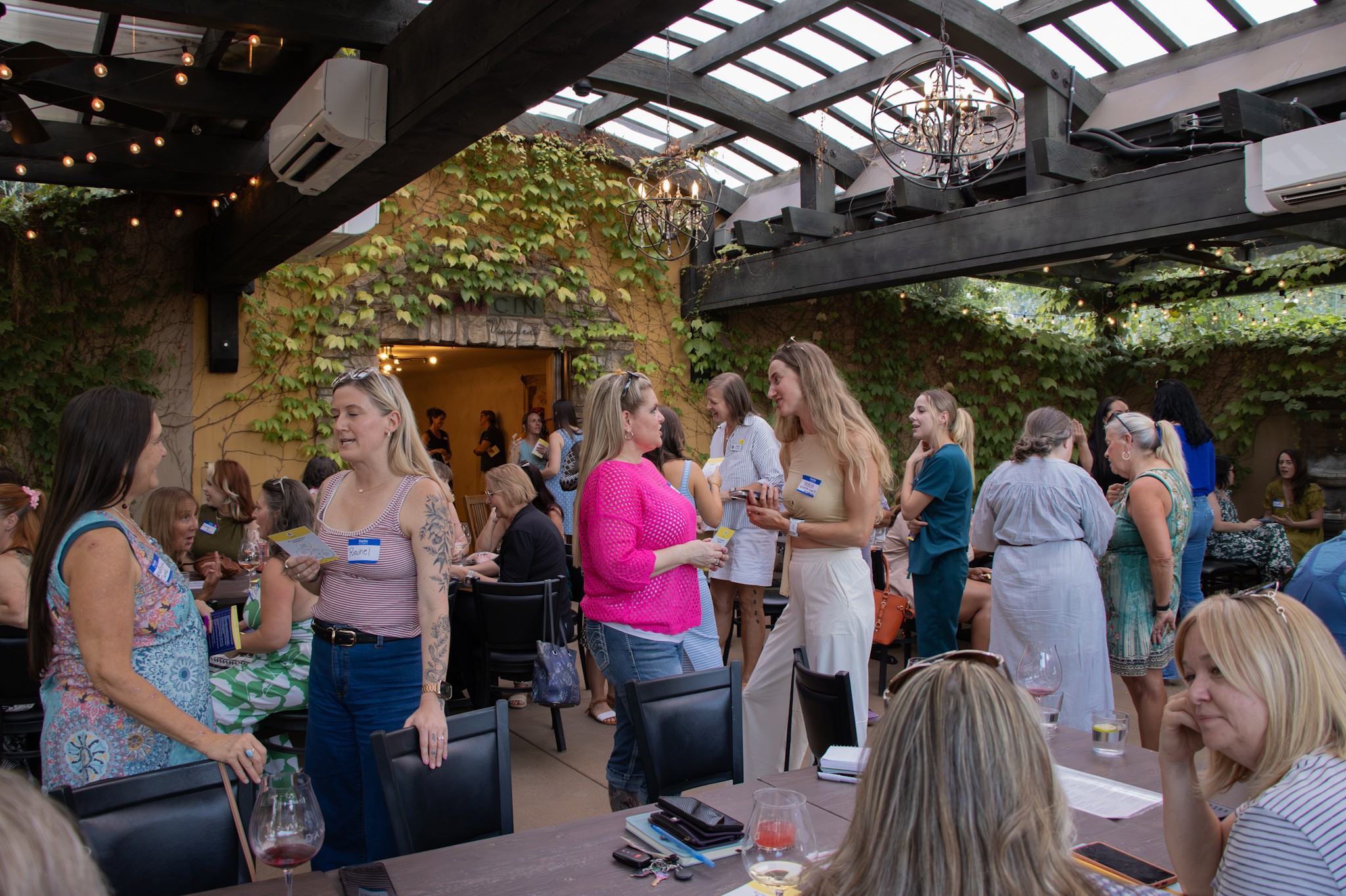 People socializing at a gathering in a covered outdoor space with greenery and string lights.
