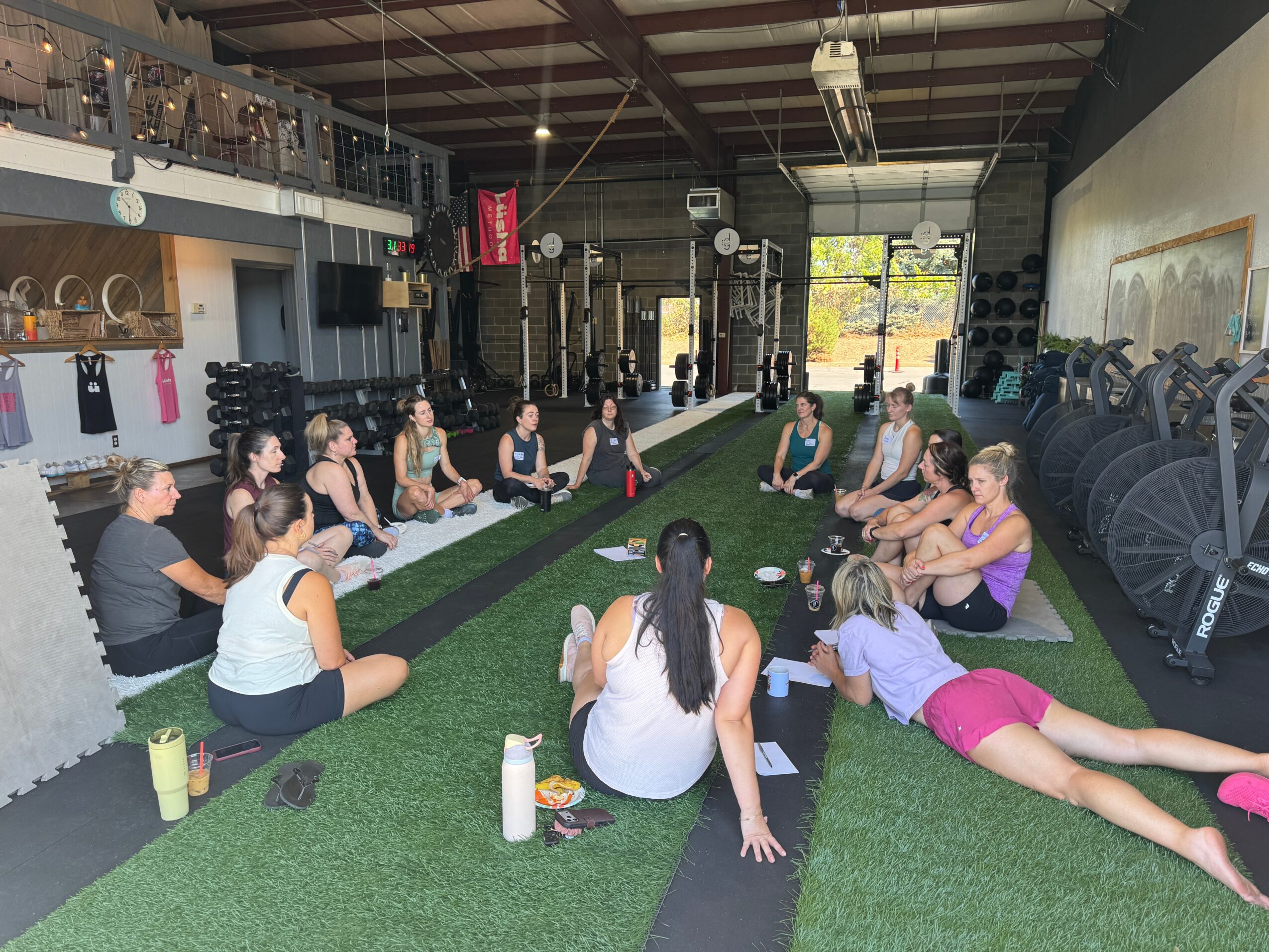 Group of people sitting and lying on mats in a gym, participating in a fitness class or workshop, with equipment and large windows in the background.