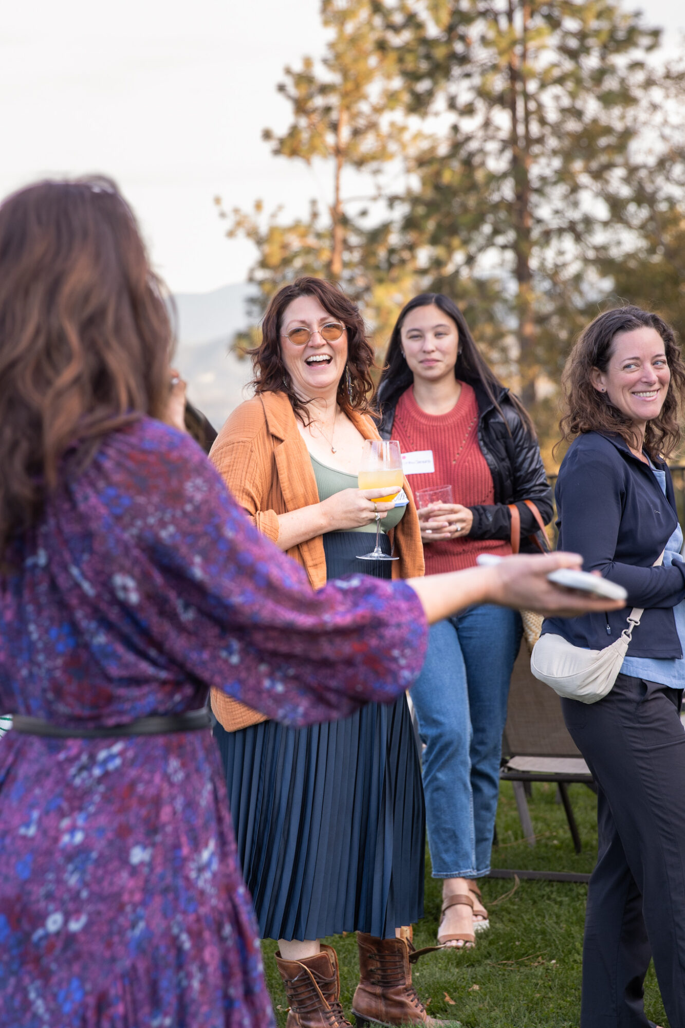 Four women outdoors, smiling and talking, with trees and sky in background.