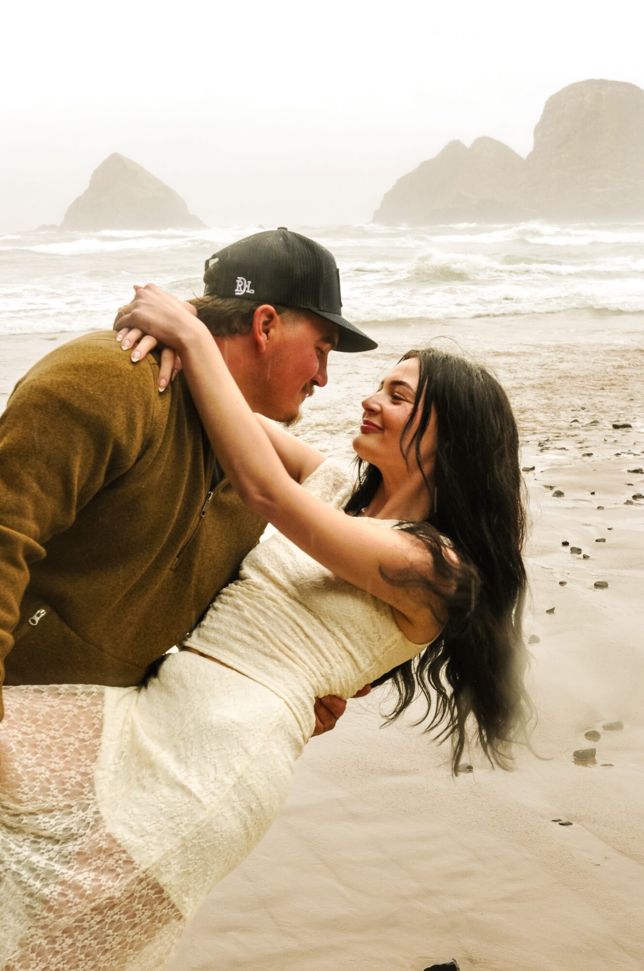A couple on a beach, smiling and embracing, with rocky formations in the background.