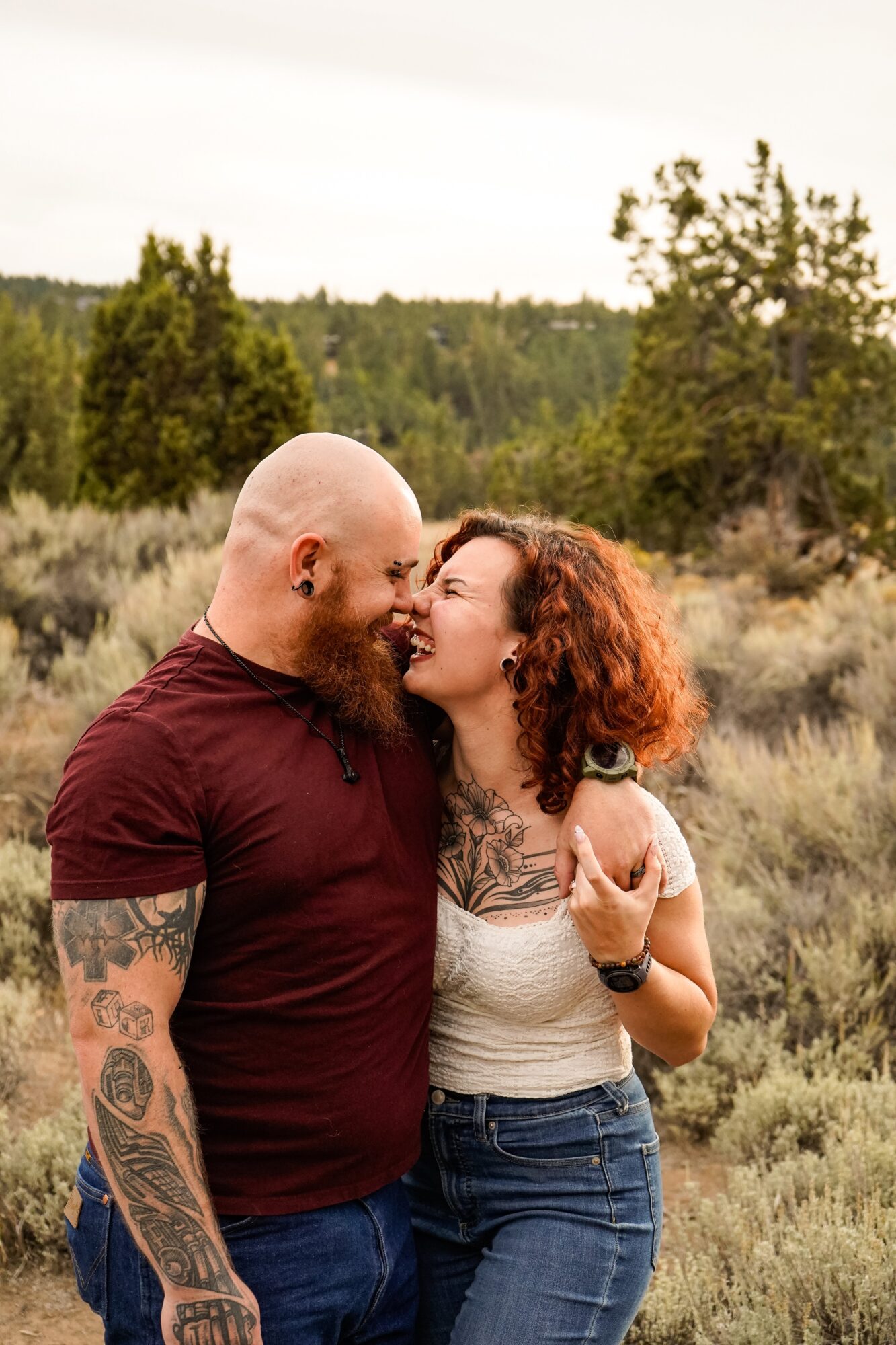 A couple stands close outdoors, faces touching, in a natural landscape with trees and grass, during daytime.