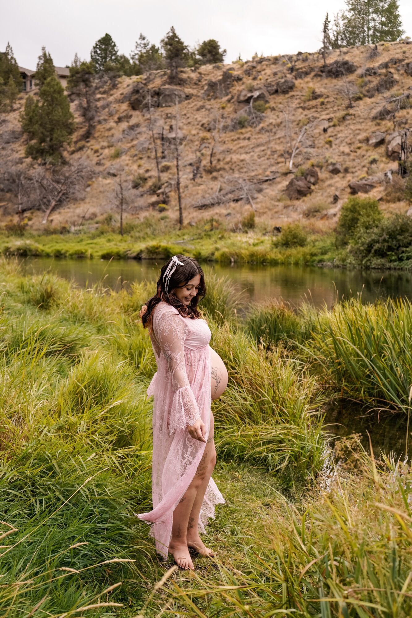 Pregnant woman in a pink dress standing in grassy area near a river, with hills and trees in background.