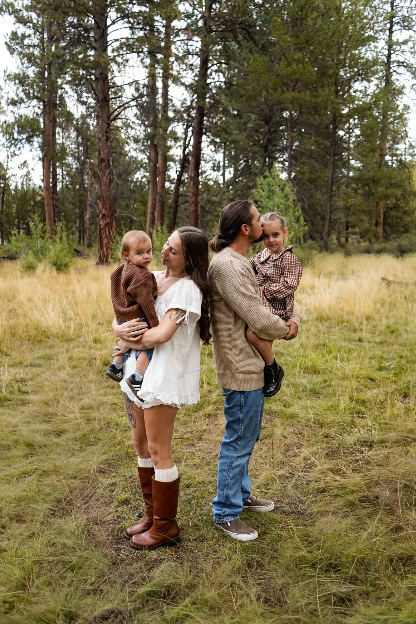 Family of four standing outdoors in a grassy field with trees, including two children and two adults, engaging with each other.