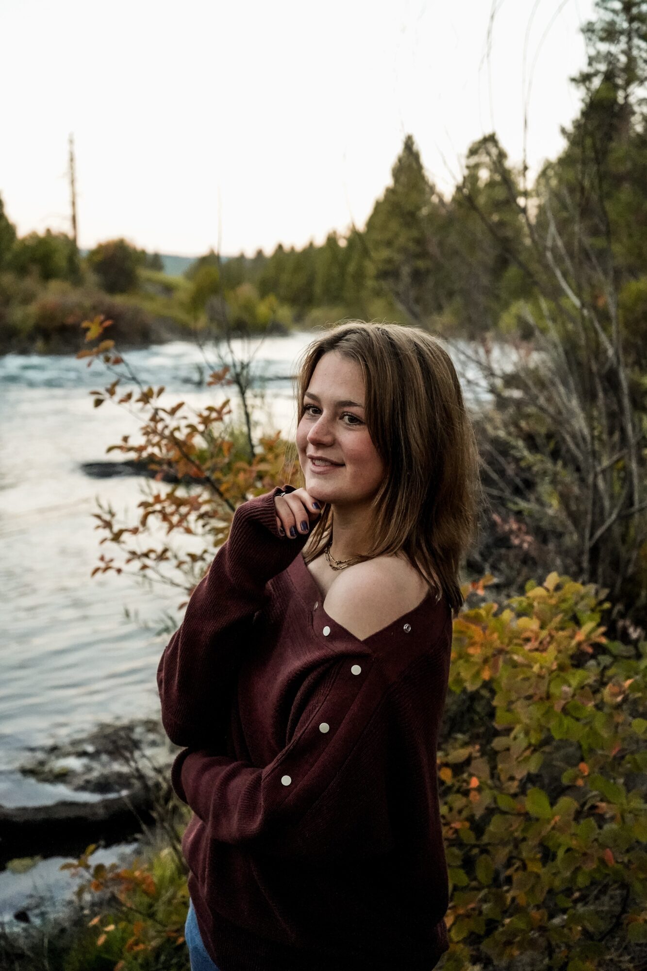 Young woman with shoulder-length hair smiling outdoors near a river, surrounded by trees and foliage.