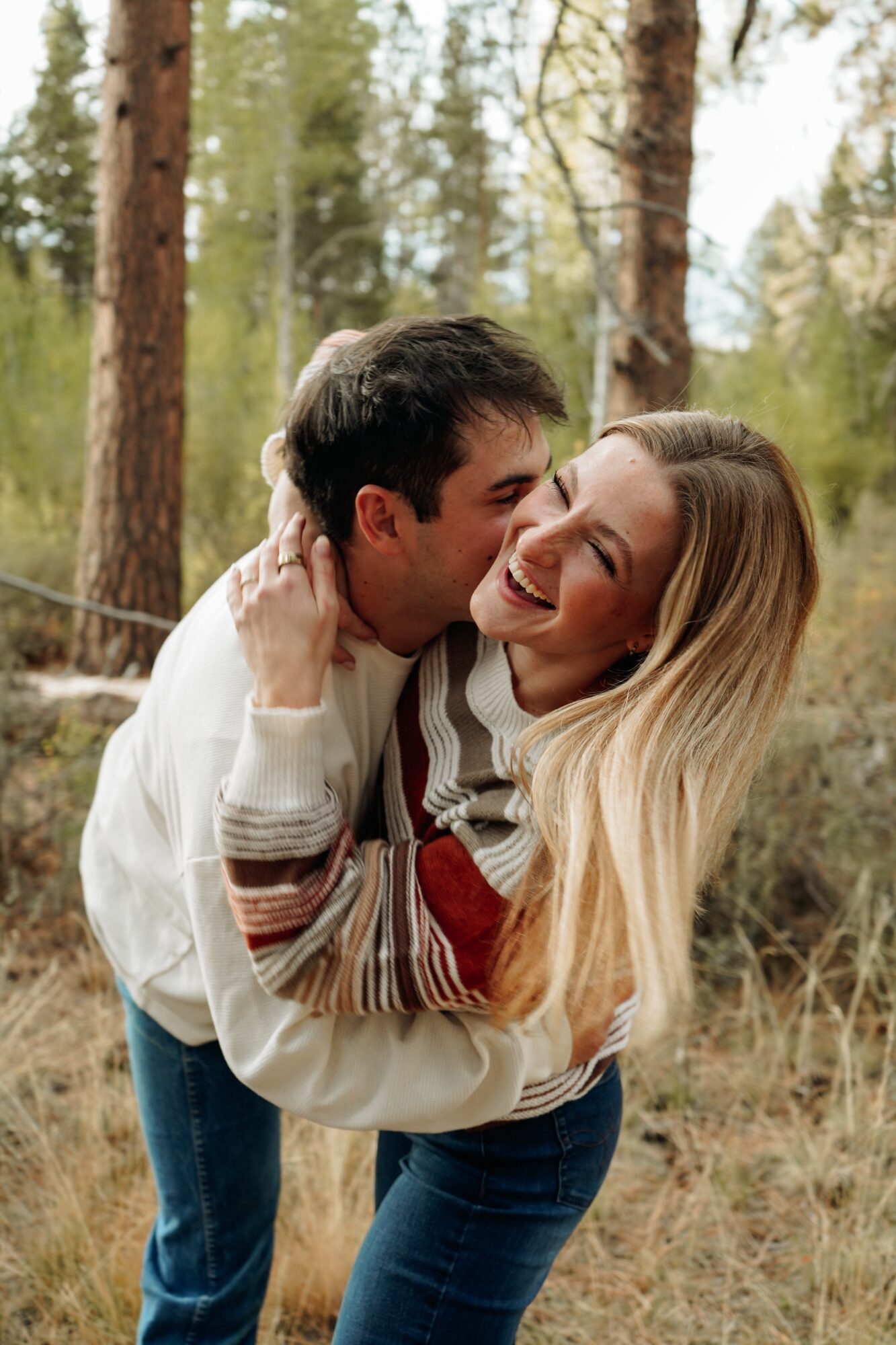 A smiling couple hugging outdoors in a forest setting, with trees and greenery in the background.