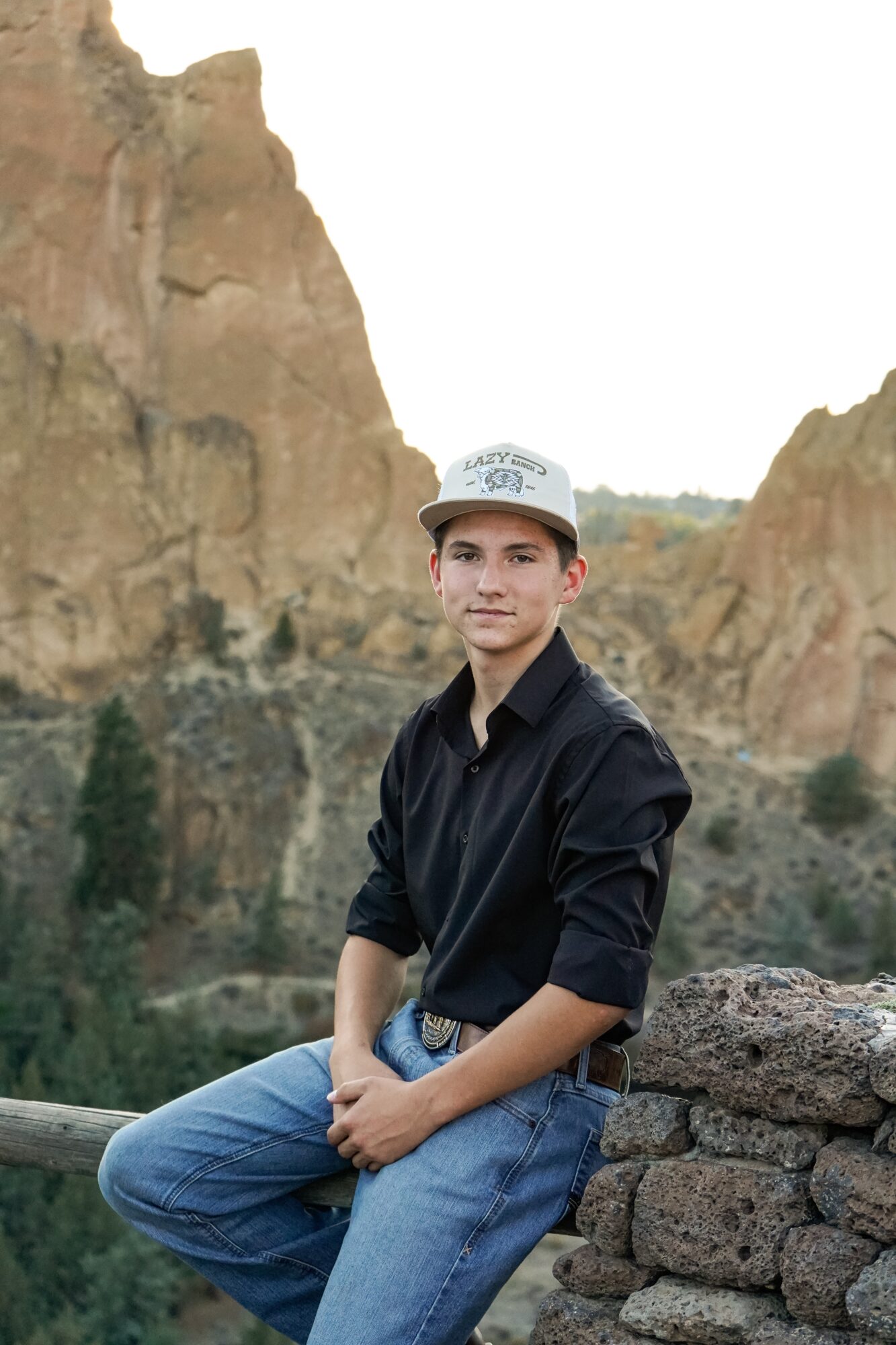 Young person sitting outdoors with rocky cliffs in background, wearing a cap, black jacket, and jeans.