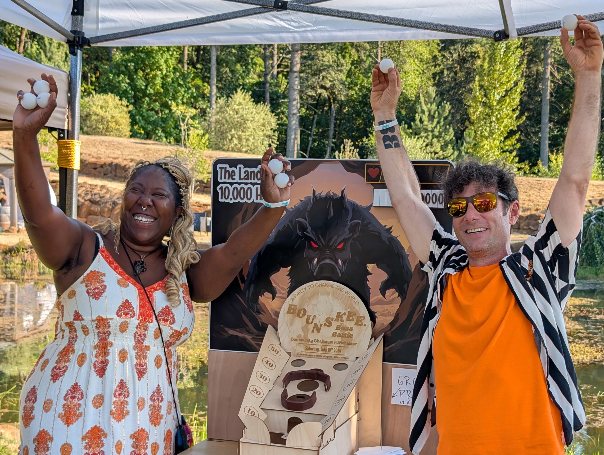 Two people celebrate outdoors under a canopy, raising their fists, with a sign and a gorilla image in the background.
