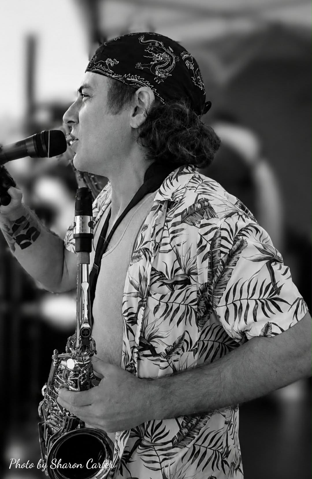 Person with curly hair, wearing a bandana and floral shirt, playing a saxophone and singing into a microphone.