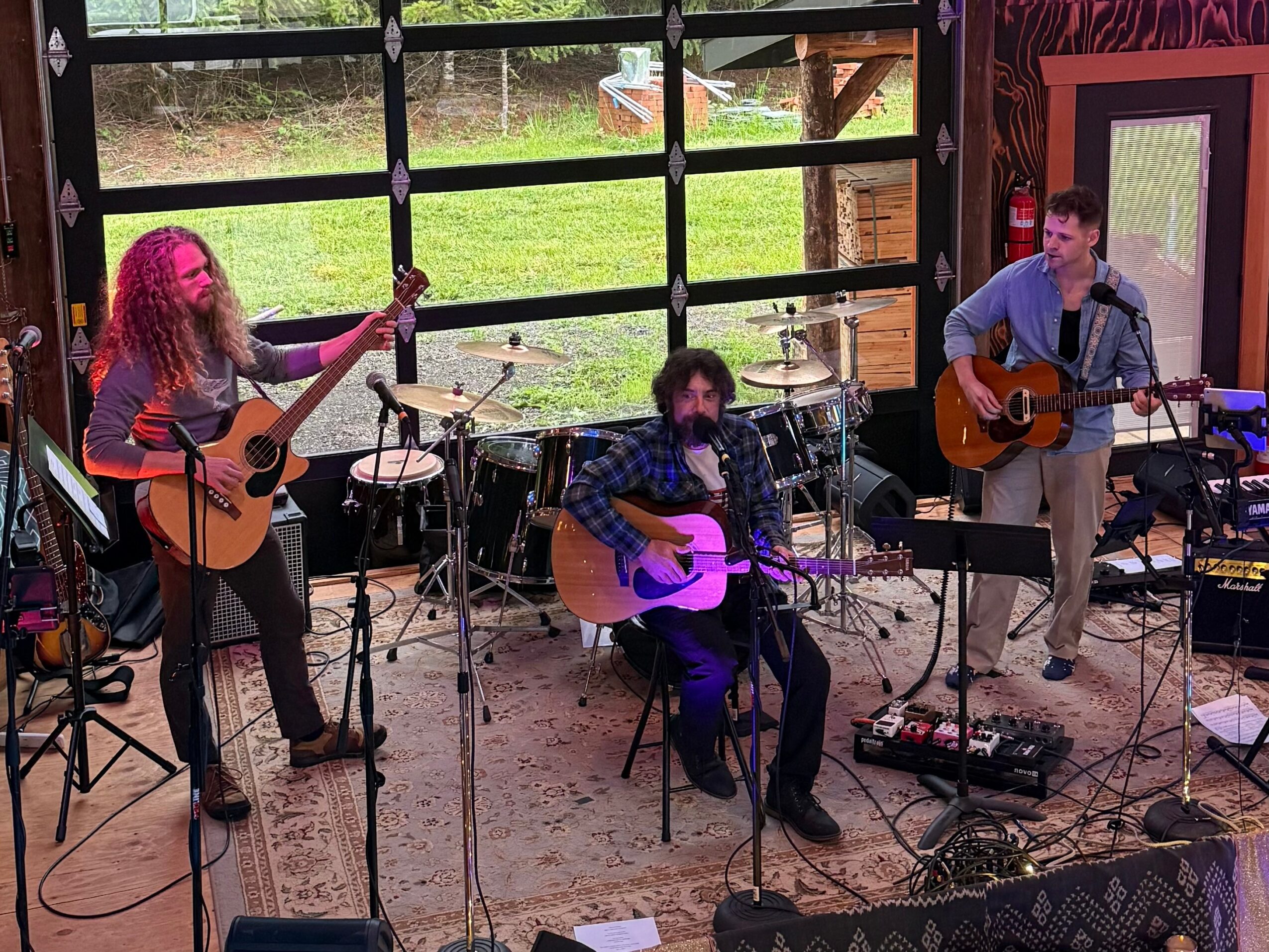Three musicians perform with guitars and drums in a room with large windows and colorful walls.