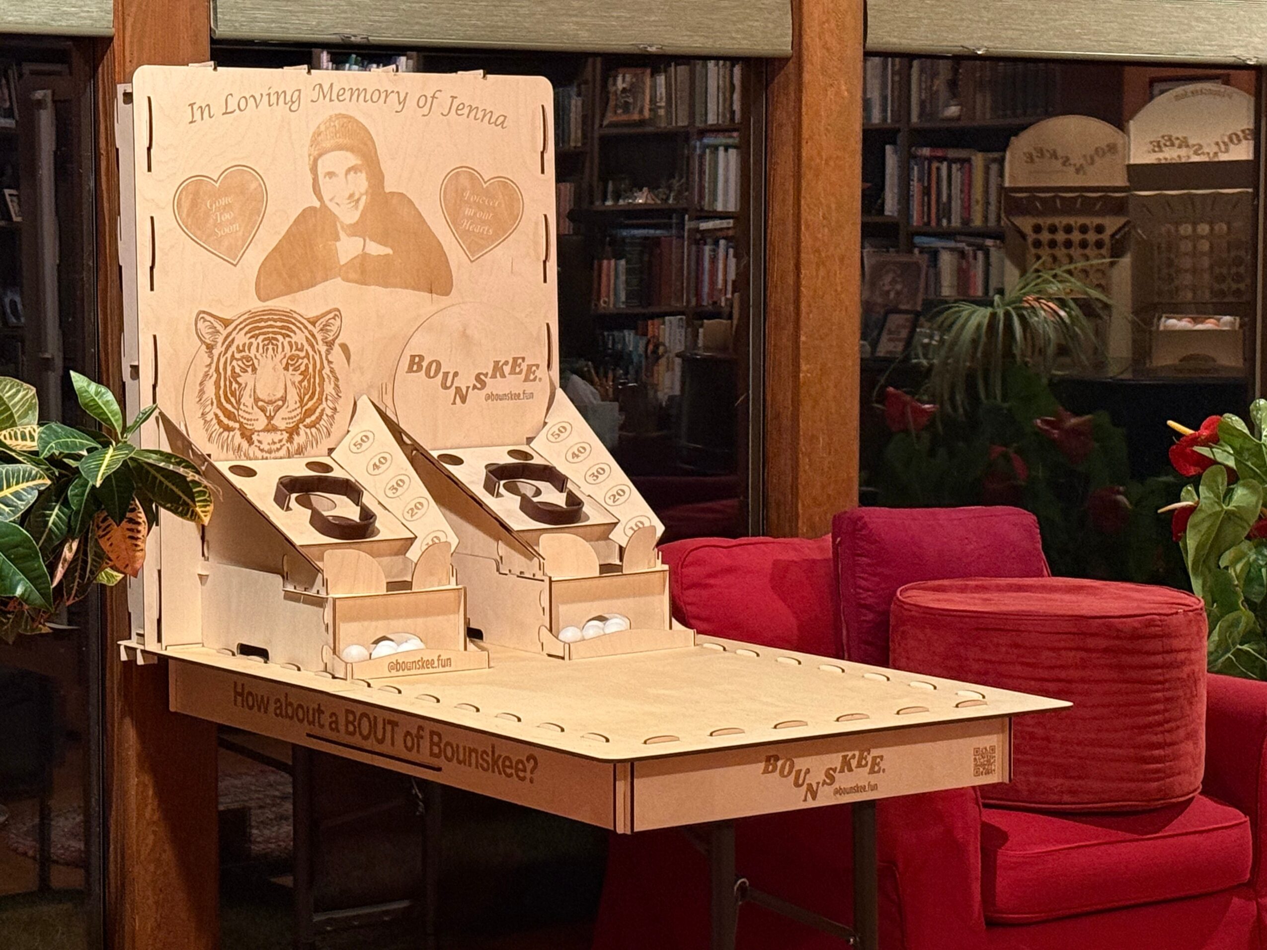 Book display with a portrait, hearts, tiger, and text, on a table with red chairs and plants in a library setting.
