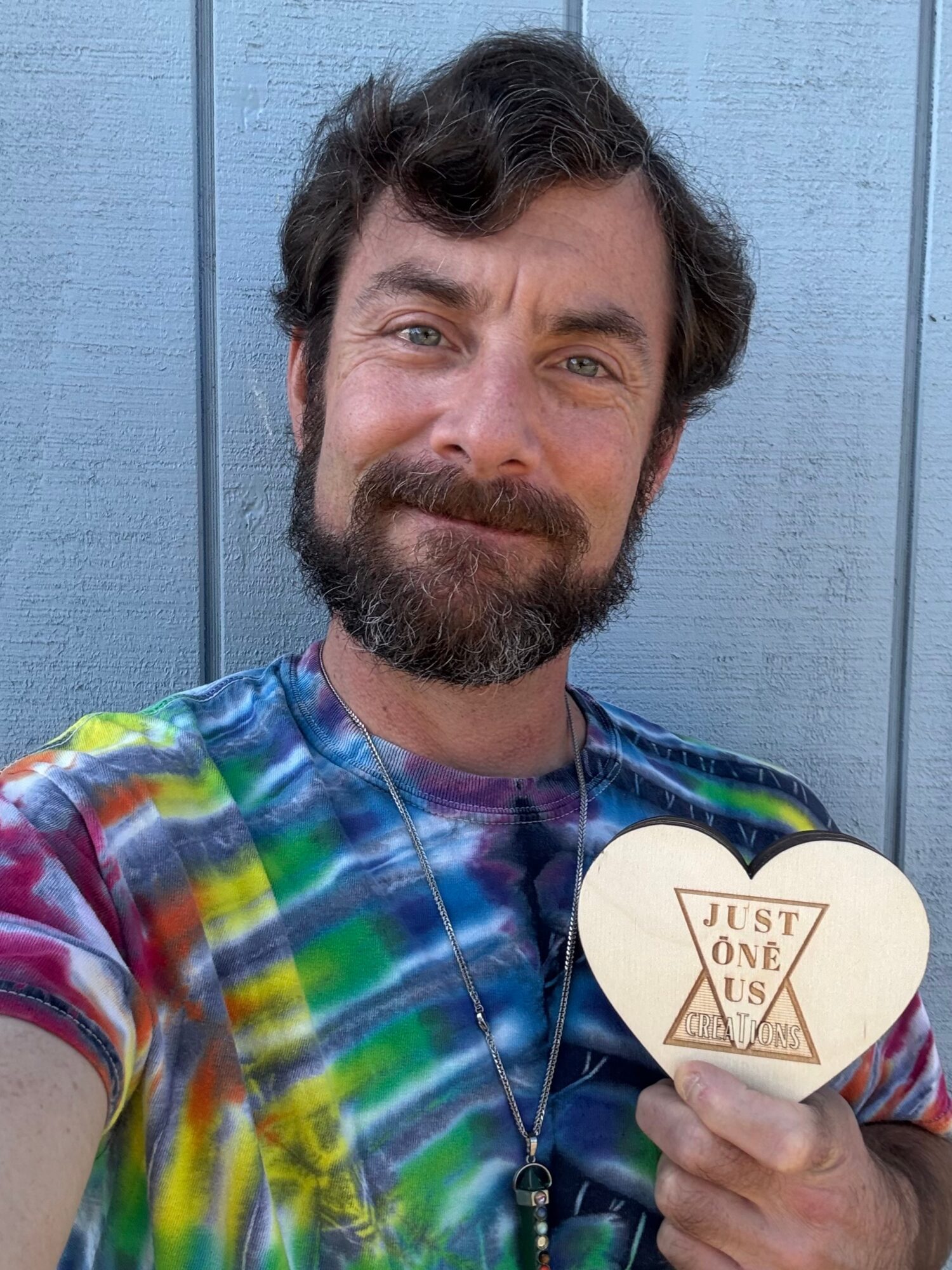Man with beard and curly hair smiling, holding a guitar pick, wearing a colorful tie-dye shirt, standing in front of a blue wall.