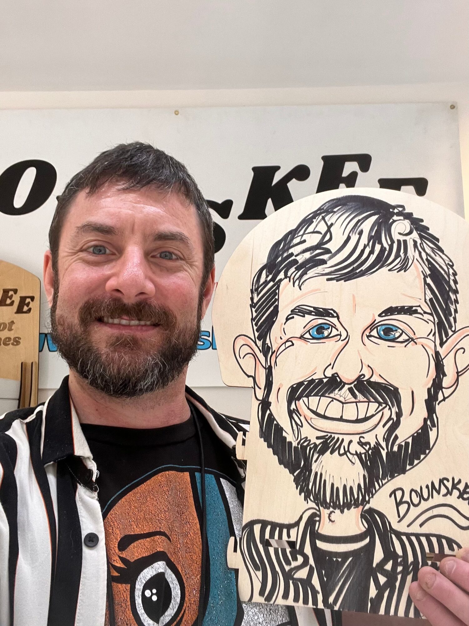 Man with dark hair and beard smiling, holding a caricature drawing of himself, in front of a sign with partially visible text.