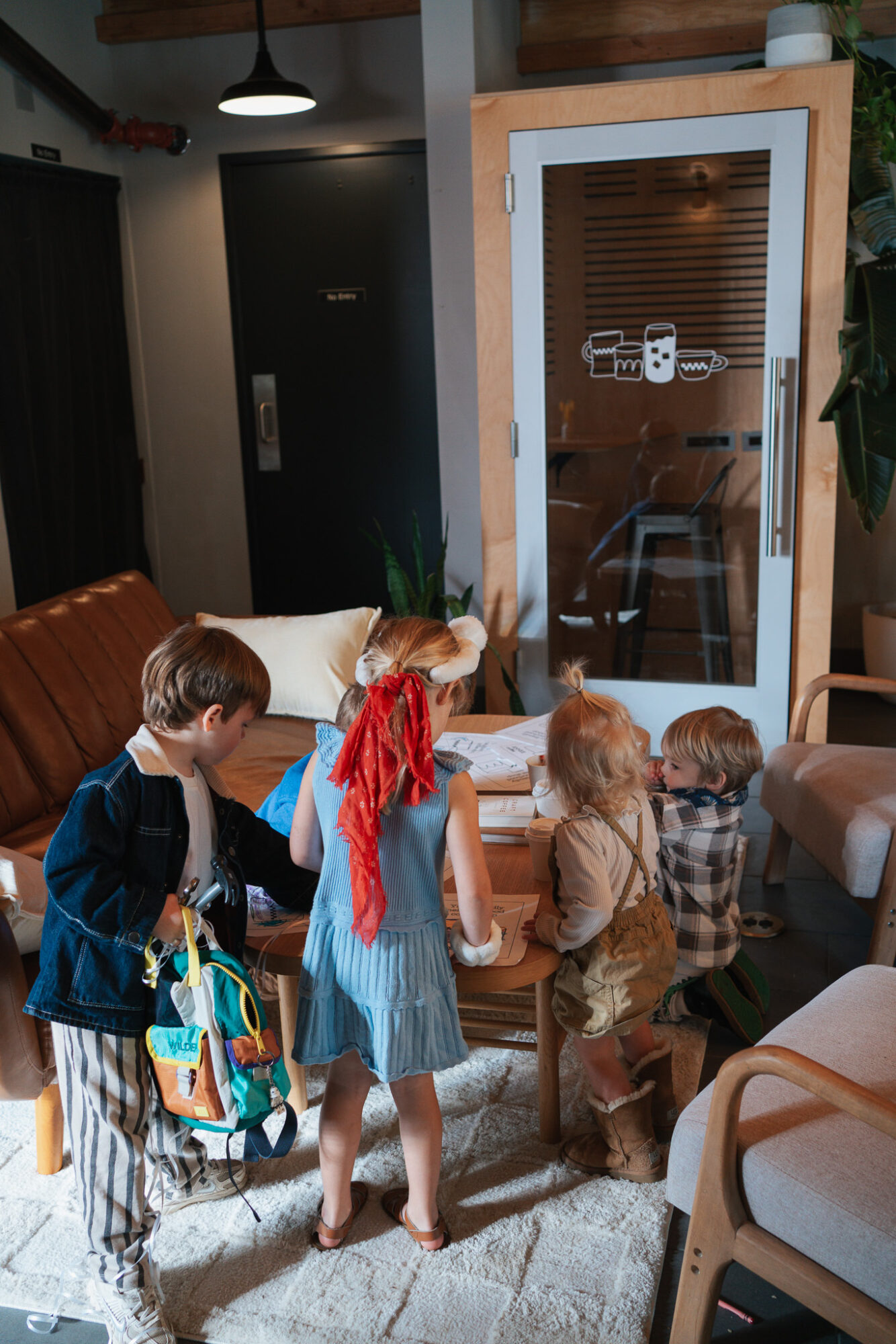 Four children gather around a small table in a cozy room, with a door and plants in the background.