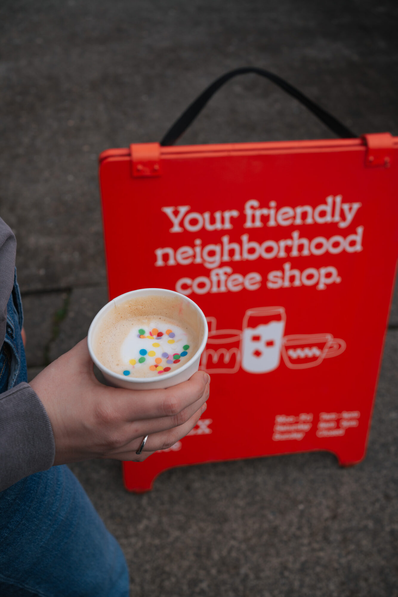 Person holding a cup of colorful cereal with a red sign advertising a neighborhood coffee shop.