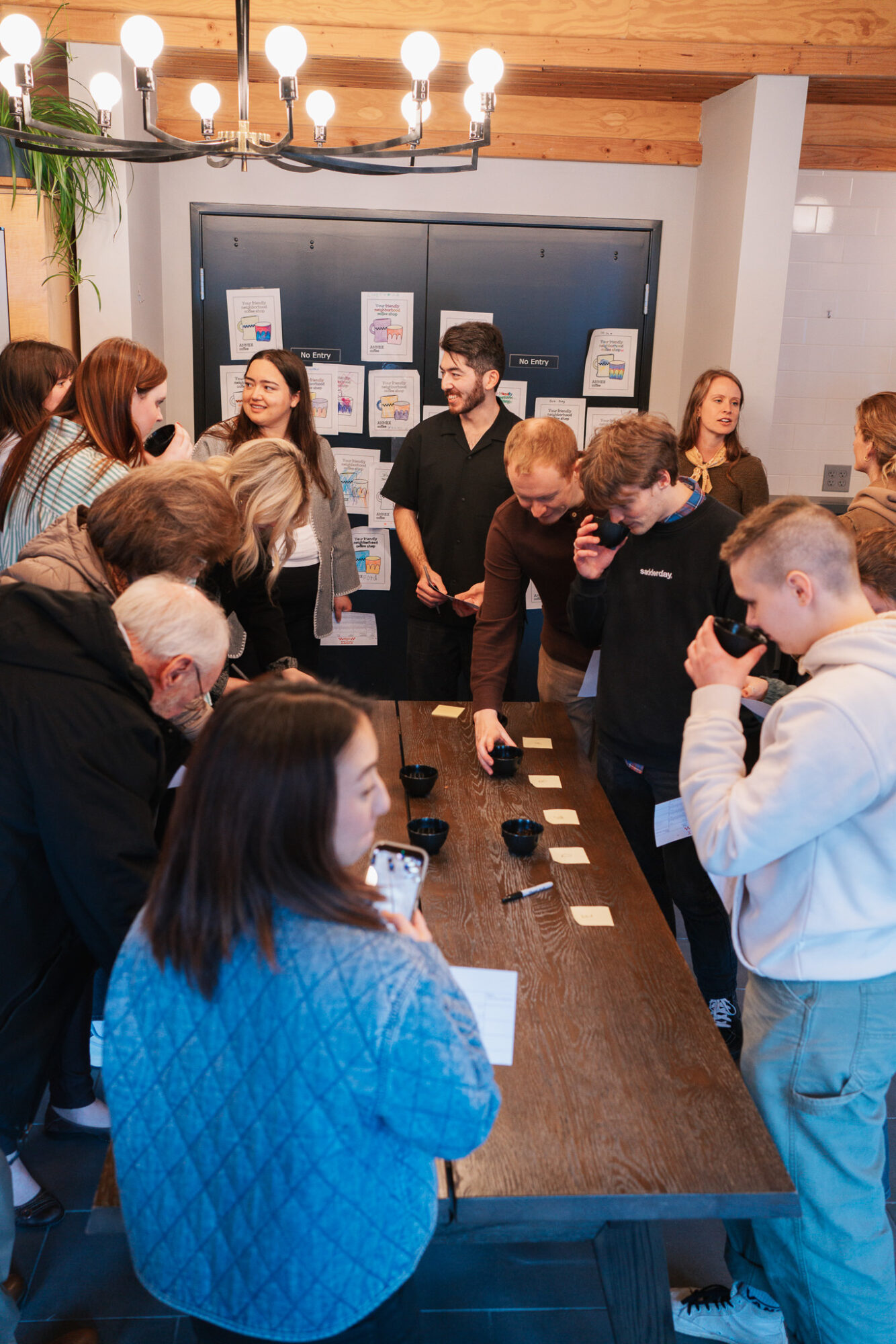 Group of people gathered around a table in a room with a blackboard and artwork, some talking and others observing.
